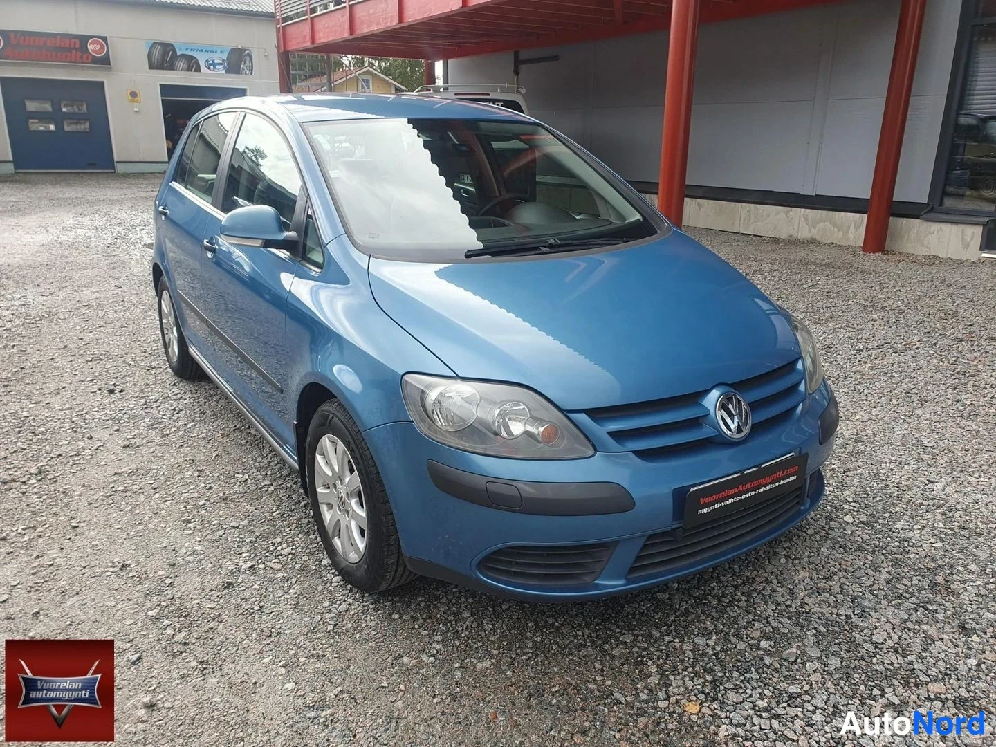 Etuviisto, nokka oikealle: Sininen Volkswagen Golf Plus etukolmasosalta, nokka oikealla, auton vasen kylki ja etuosa näkyvät sorapihalla myyntihallin edustalla.