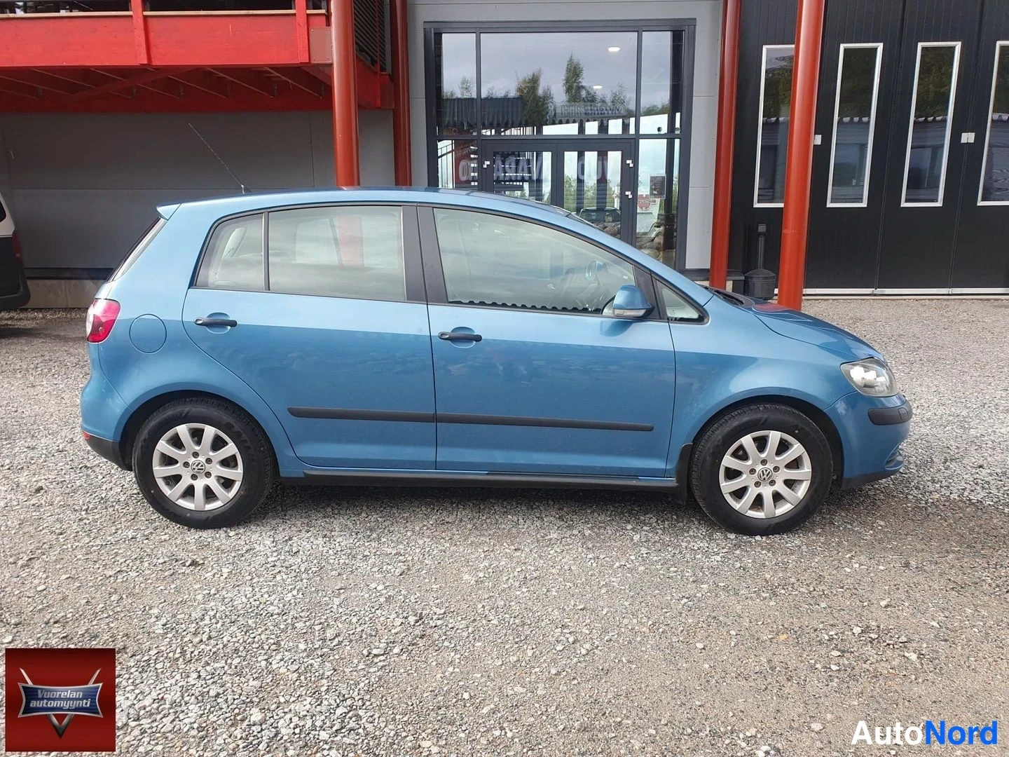 Etuviisto, nokka oikealle: Vaaleansininen Volkswagen Golf Plus kuvattuna auton vasemmalta etukulmasta, keula oikealla; auto seisoo sorapohjalla punaisen rakennuksen edessä.