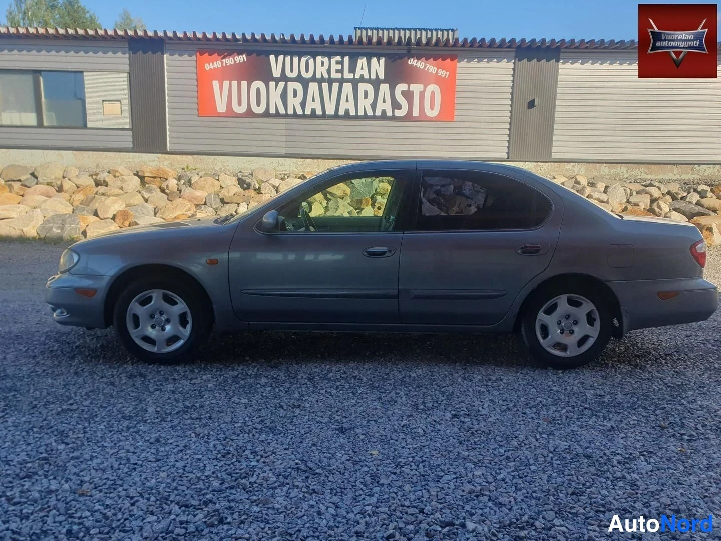 Etuviisto, nokka vasemmalle: Nissan Maxima -sedan profiilikuva nokka vasemmalla sorapinnalla; keulan ajovalo ja takavalojen reunat erottuvat, taustalla kivilatomus ja harmaa varast