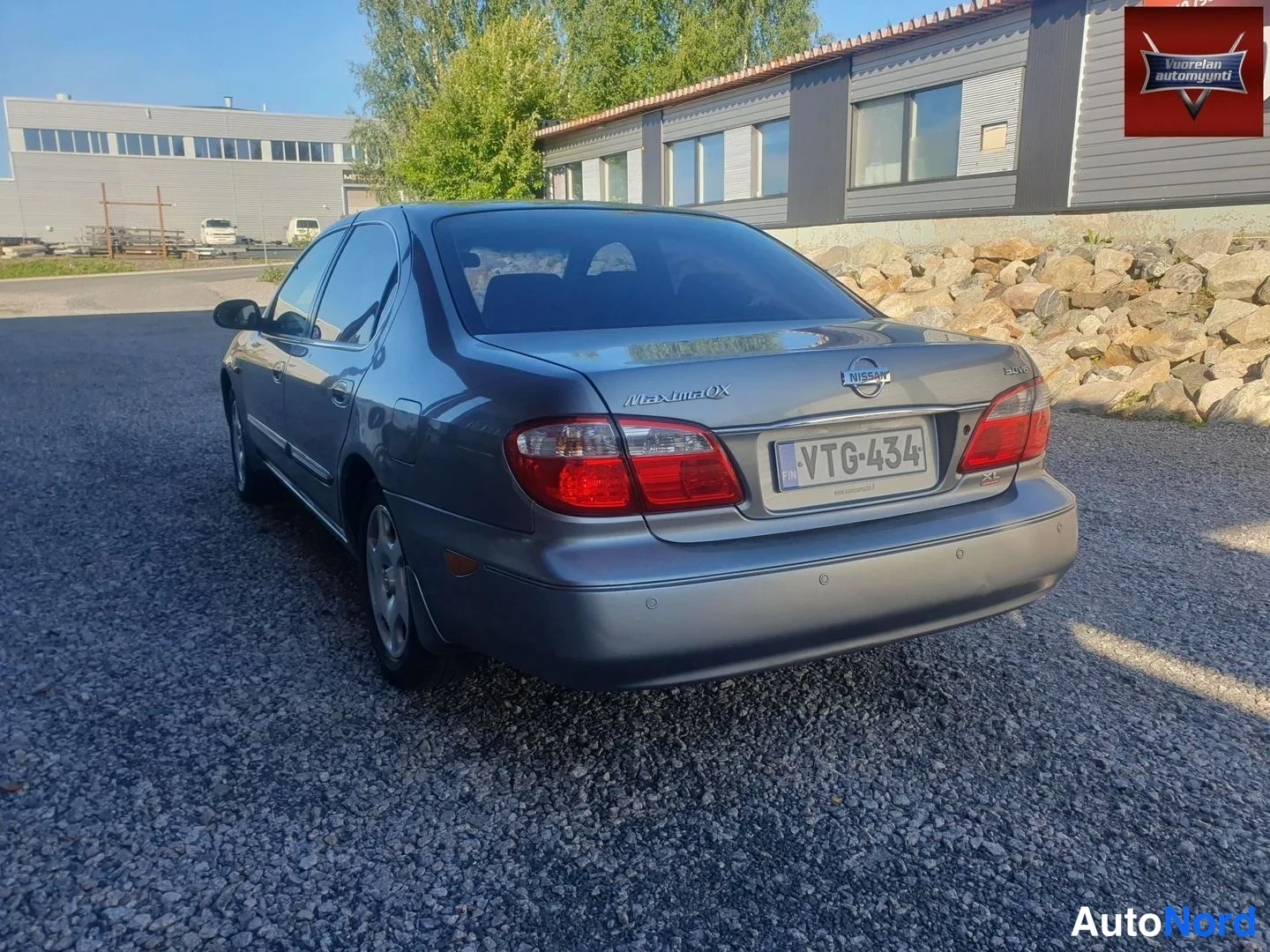Takaviisto: Auton takakolmannes: hopeanharmaa sedan, rekisterikilpi, perässä Maxima QX -teksti ja Nissan-merkki.