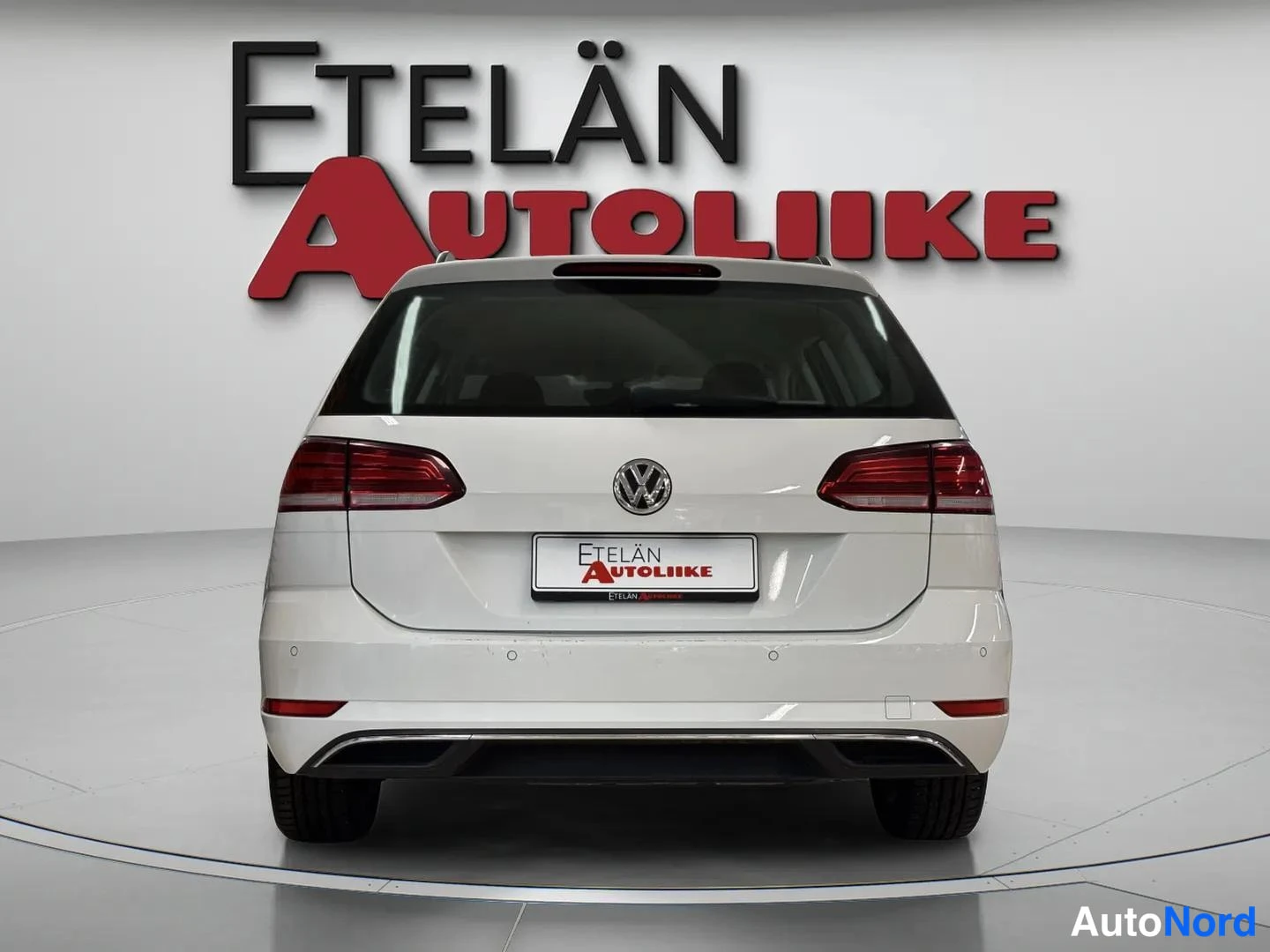 Takaa: Valkoisen farmariauton suora takanäkymä: VW-tunnus, takavalot, rekisterikilven paikalla mainoslaatta ja takapuskuri.
