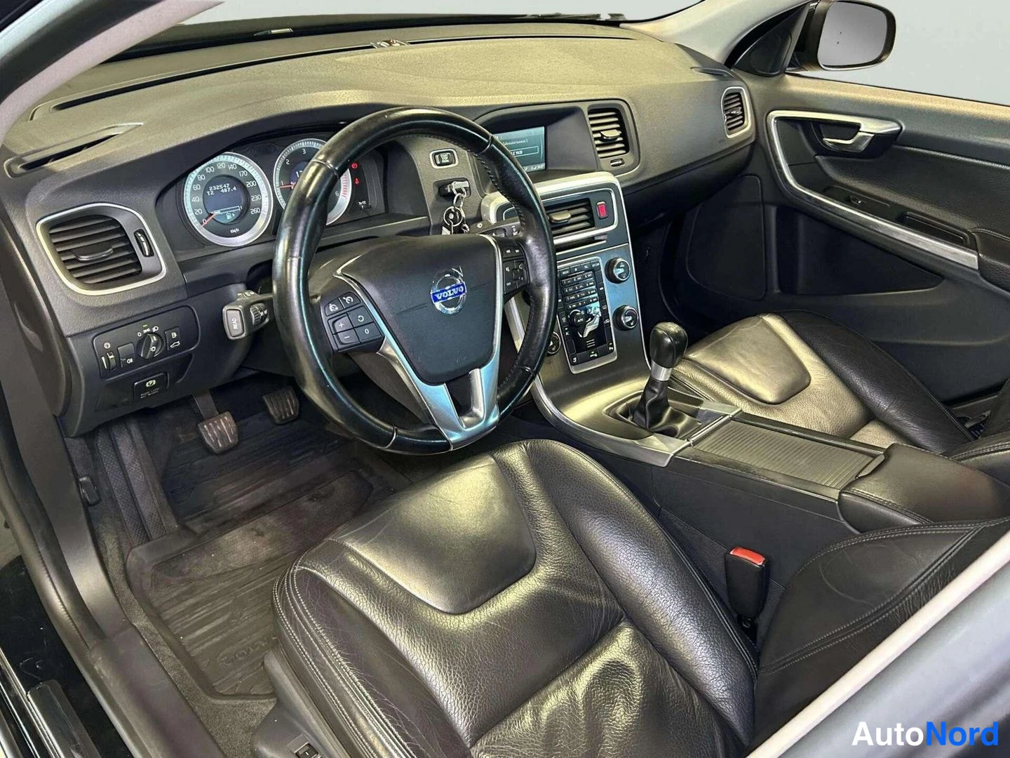 Ohjaamo: Interior front cabin view showing driver's side steering wheel with multifunction controls, instrument cluster, gear selector lever and black leather seat.