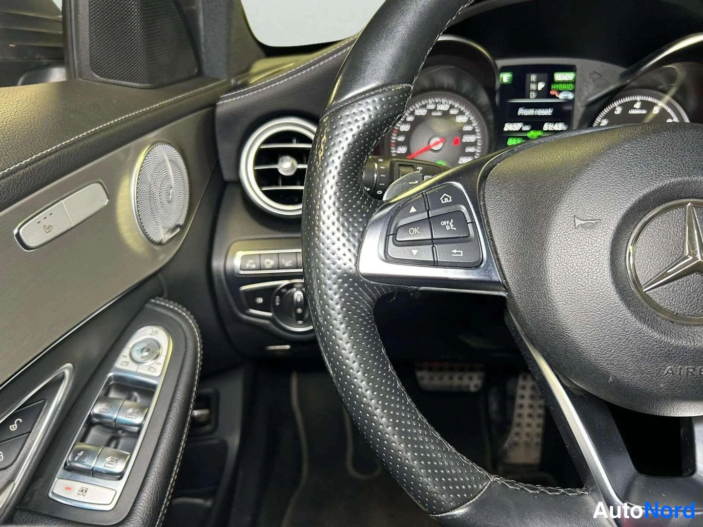 Edestä: Sisäkuva auton ohjaamosta: musta Mercedes-Benz-ohjauspyörä, joka on kuvattu läheltä, ovipanelin ikkunannostinnapit ja kojelaudan tieto näkyvät.