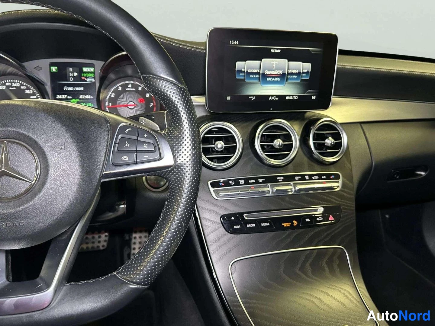 Edestä: Sisäkuva kojelaudasta: Mercedes-Benz-ohjauspyörä vasemmalla, keskellä näyttö jolla radioviestintä ja kolme pyöreää ilmastointisuuloketta.
