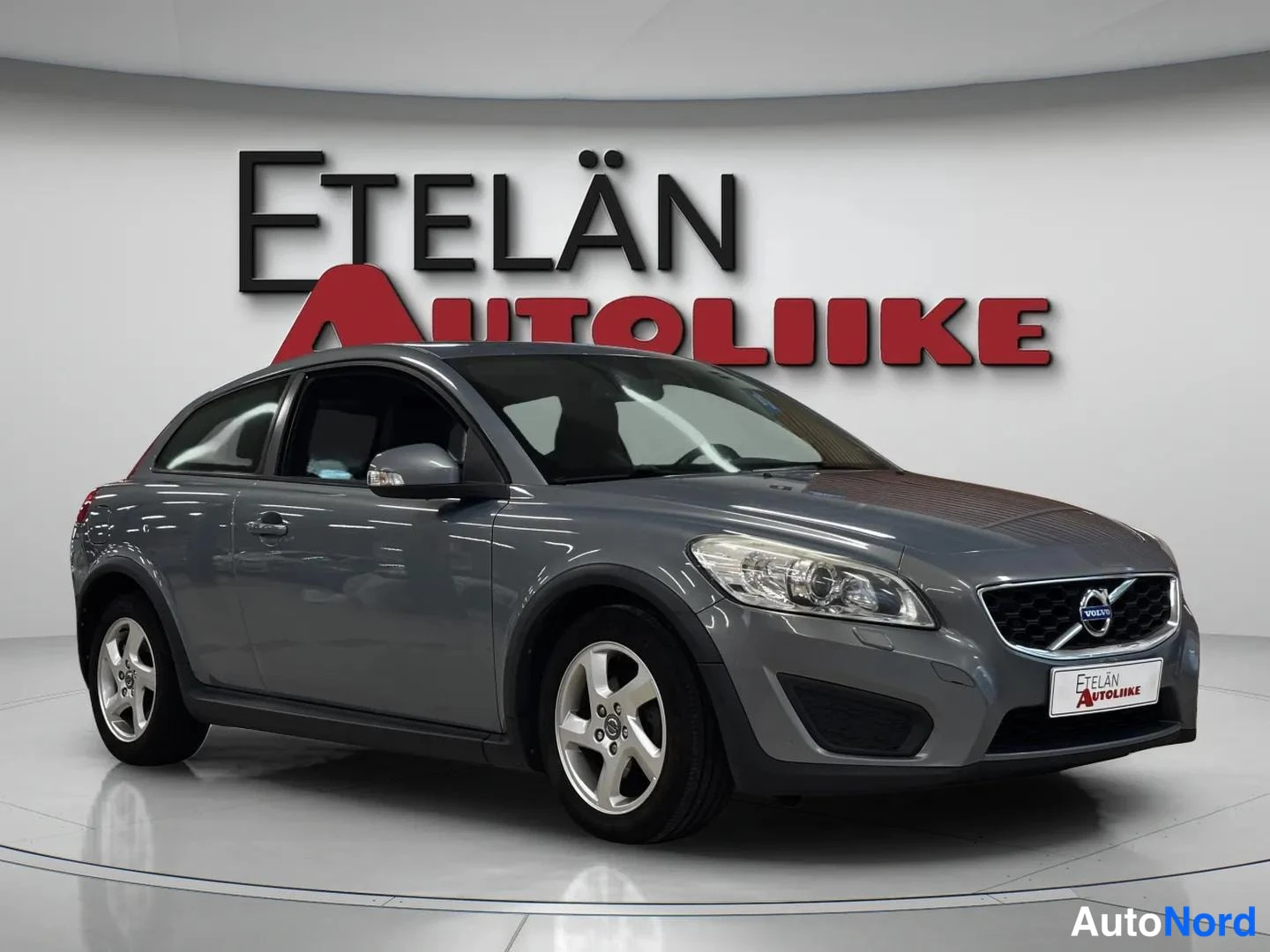 Etuviisto, nokka oikealle: Volvo C30, harmaanmetallinen coupé kuvattu kolmasosakulmasta etuosaa ja oikeaa sivua näyttäen, studiotaustalla Etelän Autoliike-logon edessä.