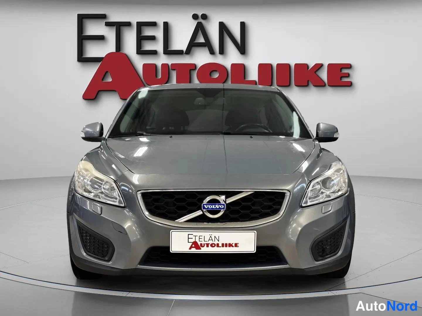 Edestä: Harmaanmetallisen Volvon etunäkymä, Volvo-merkki säleikössä ja Etelän Autoliike -kyltti rekisterikilven paikalla.