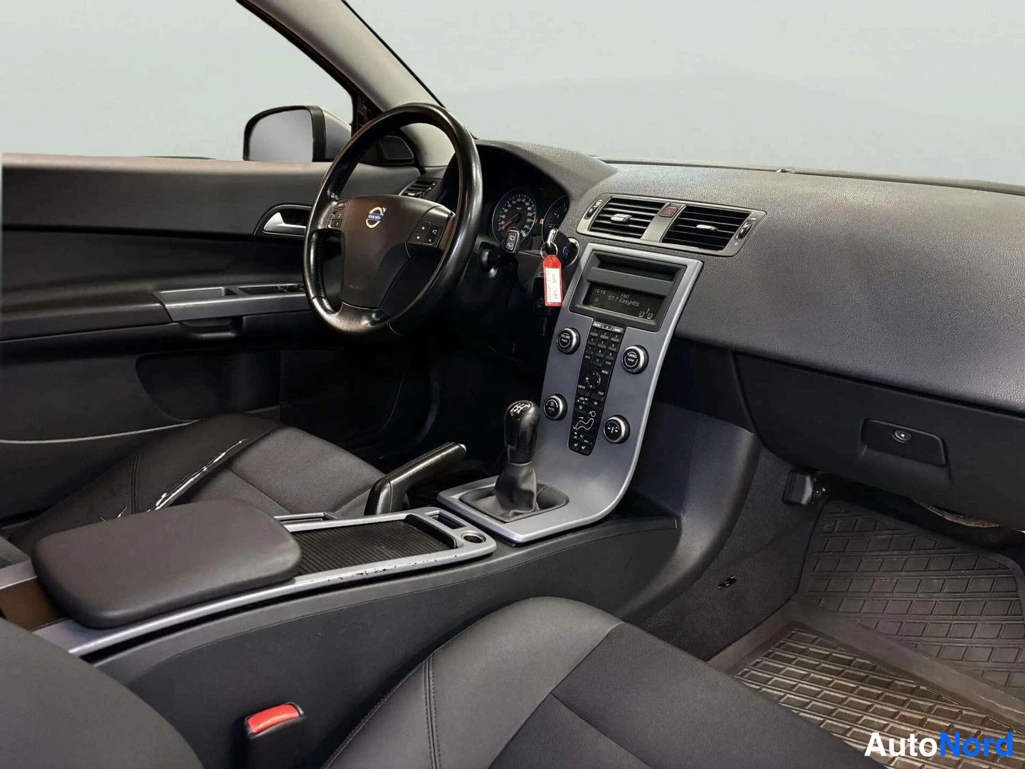 Ohjaamo: A wide view of a vehicle's interior focusing on the silver-trimmed centre console, steering wheel, dashboard vents, and front seats. The gear stick is clearly v