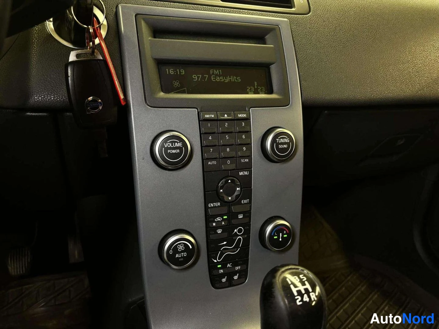 Ohjaamo: Center stack of the car interior showing a radio and climate control panel with a digital display reading 'FM1 97.7 EasyHits' and temperature '23', alongside bu