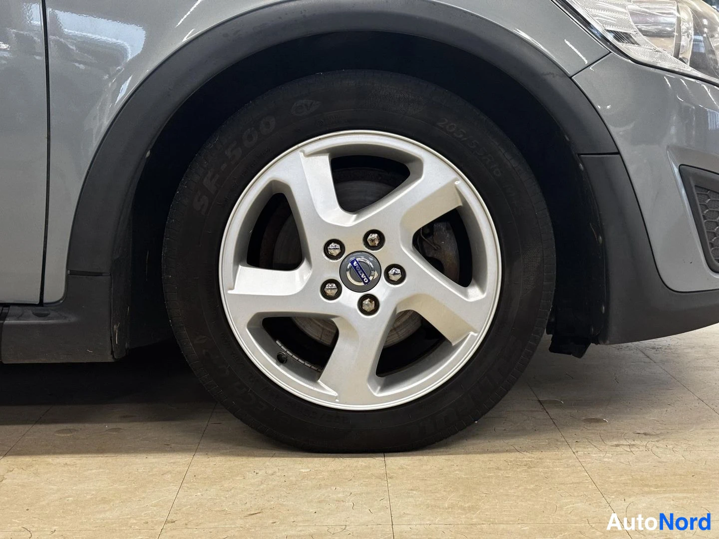 Edestä: Lähikuva auton etupyörästä: hopeanvärinen viisisakarainen Volvo-alumiinvanne, musta rengas ja harmaa kori sekä tummat lokasuojaukset.