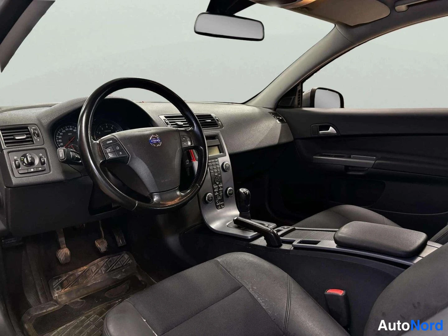 Ohjaamo: Black interior of a car showing the driver's seat, steering wheel with the Volvo logo, instrument cluster, centre console with climate controls and an audio uni