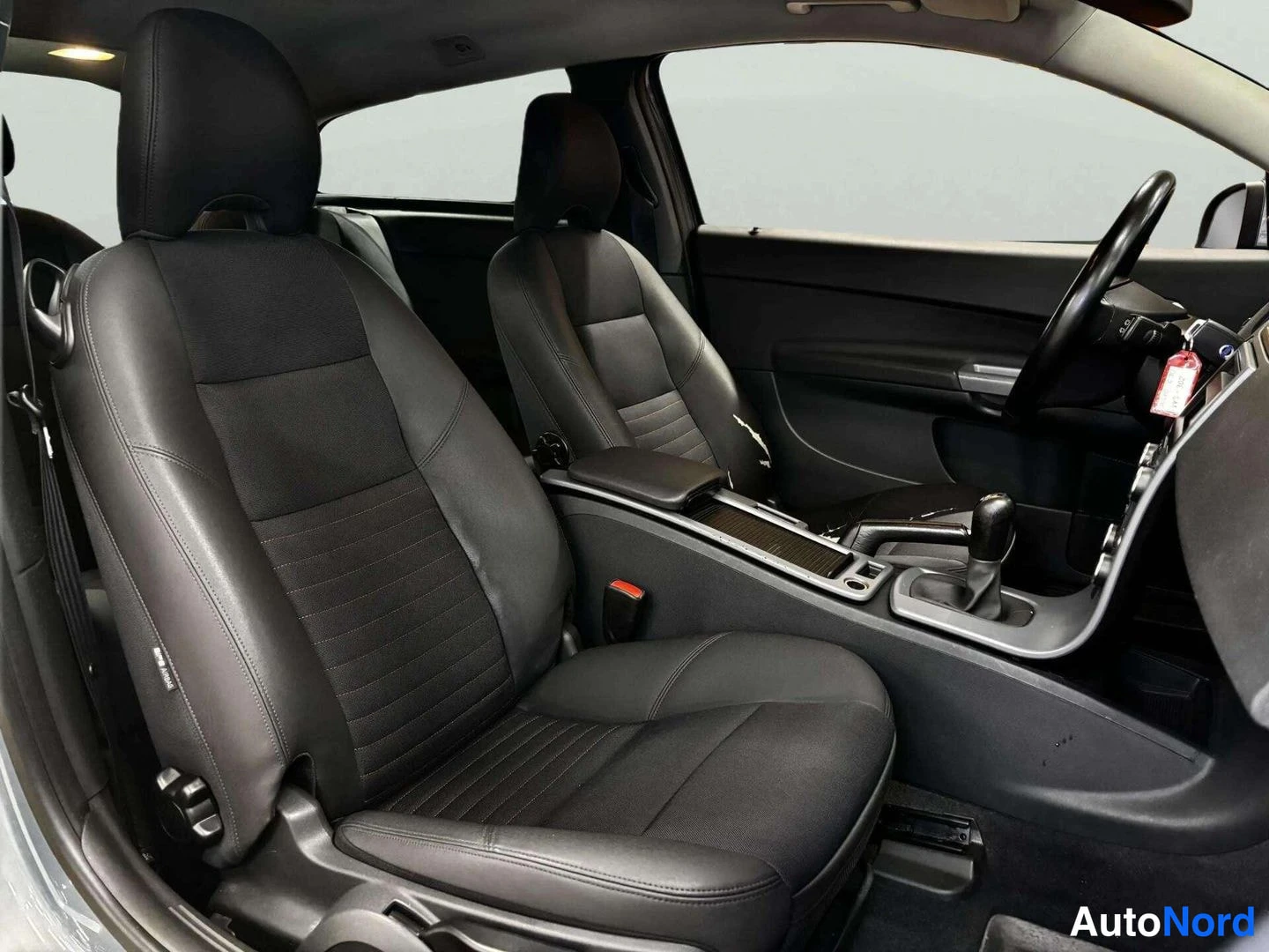 Etuistuimet: View into a black car cabin from the open passenger door, showing two front seats with a sporty design and a manual gear lever in the centre console.