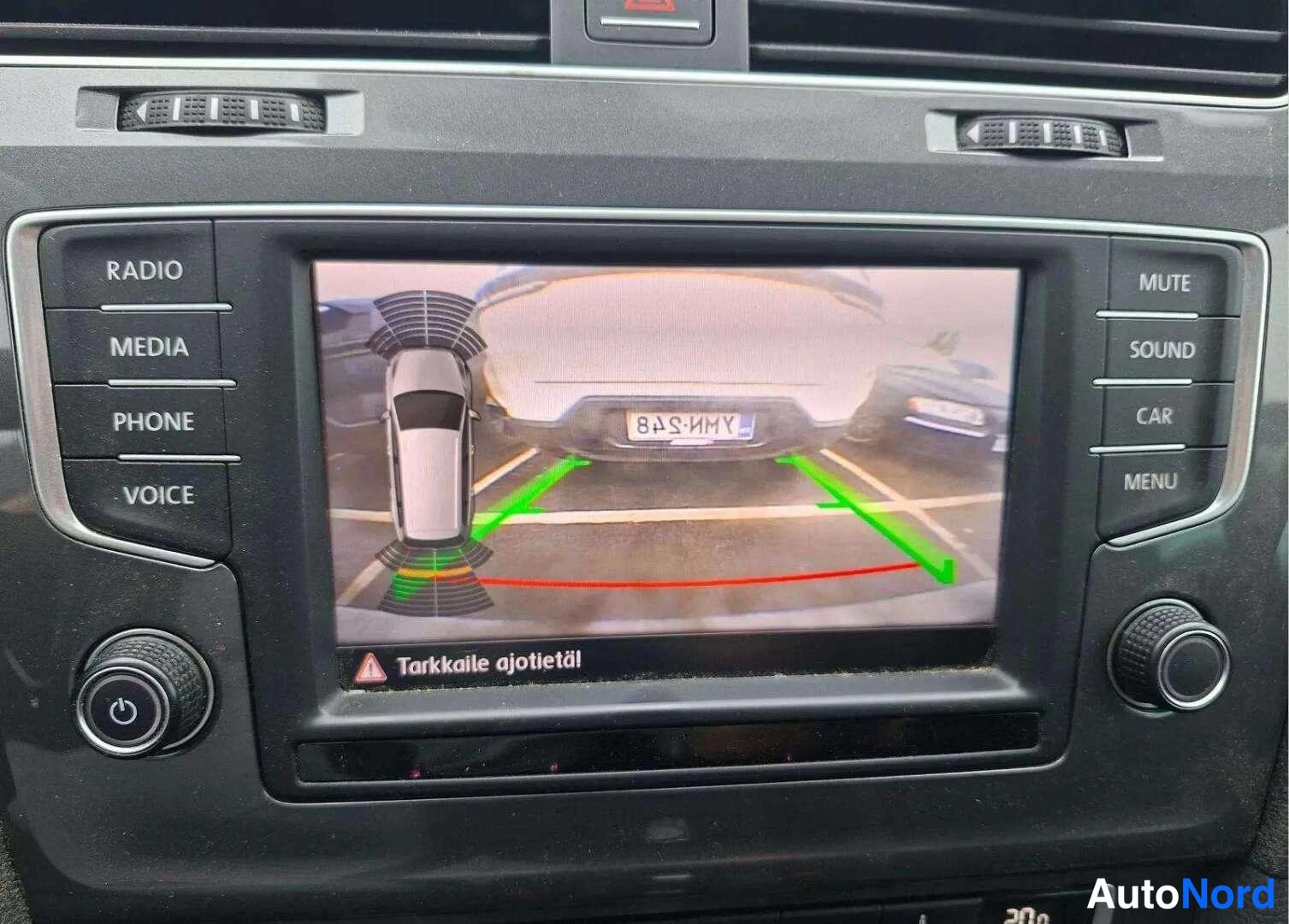 Muu: Kojelaudan infotainment-näyttö näyttää peruutuskameran kuvan pysäköintitunnustajilla ja tekstillä Tarkkaile ajetietä.