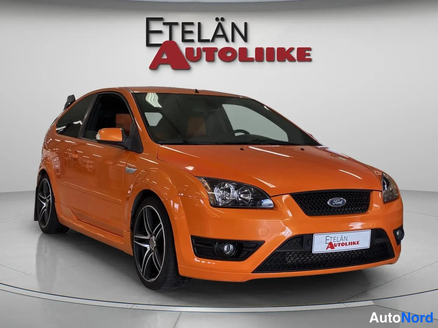 Etuviisto, nokka oikealle: Oranssi Ford-kolmiosen etu ja vasen kylki studiosta nähtynä, jossa näkyy tummansiniset ja mustat vanteet, edessa oleva logo ja Etelän Autoliike-tunnus.