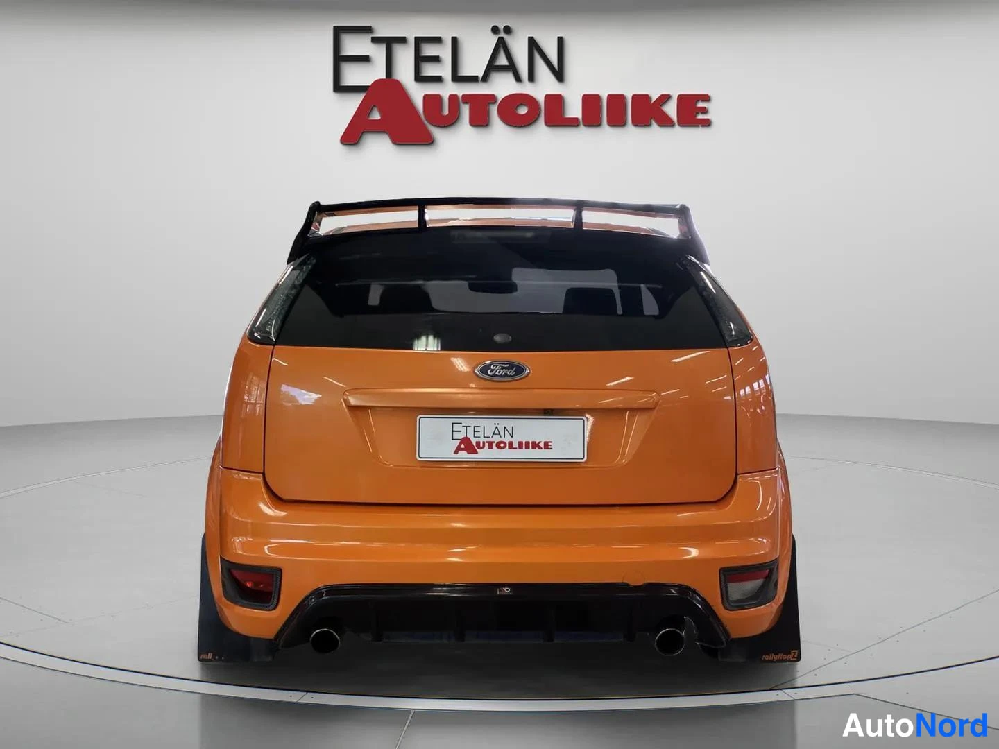 Takaa: Oranssin auton takaosa suoraan takaa; Ford-merkki, suuri kattospoileri ja kaksoispakoputket, rekisterikilven paikalla Etelän Autoliike -kyltti.