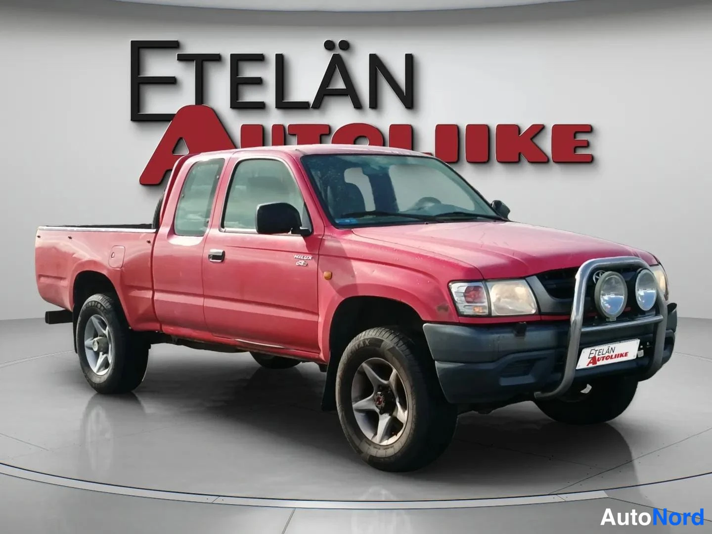 Etuviisto, nokka oikealle: Punainen Toyota Hilux -pakettiauto etuviistokuva studiossa, etupuskqueriin asennettu kromikaari ja lisävalot.