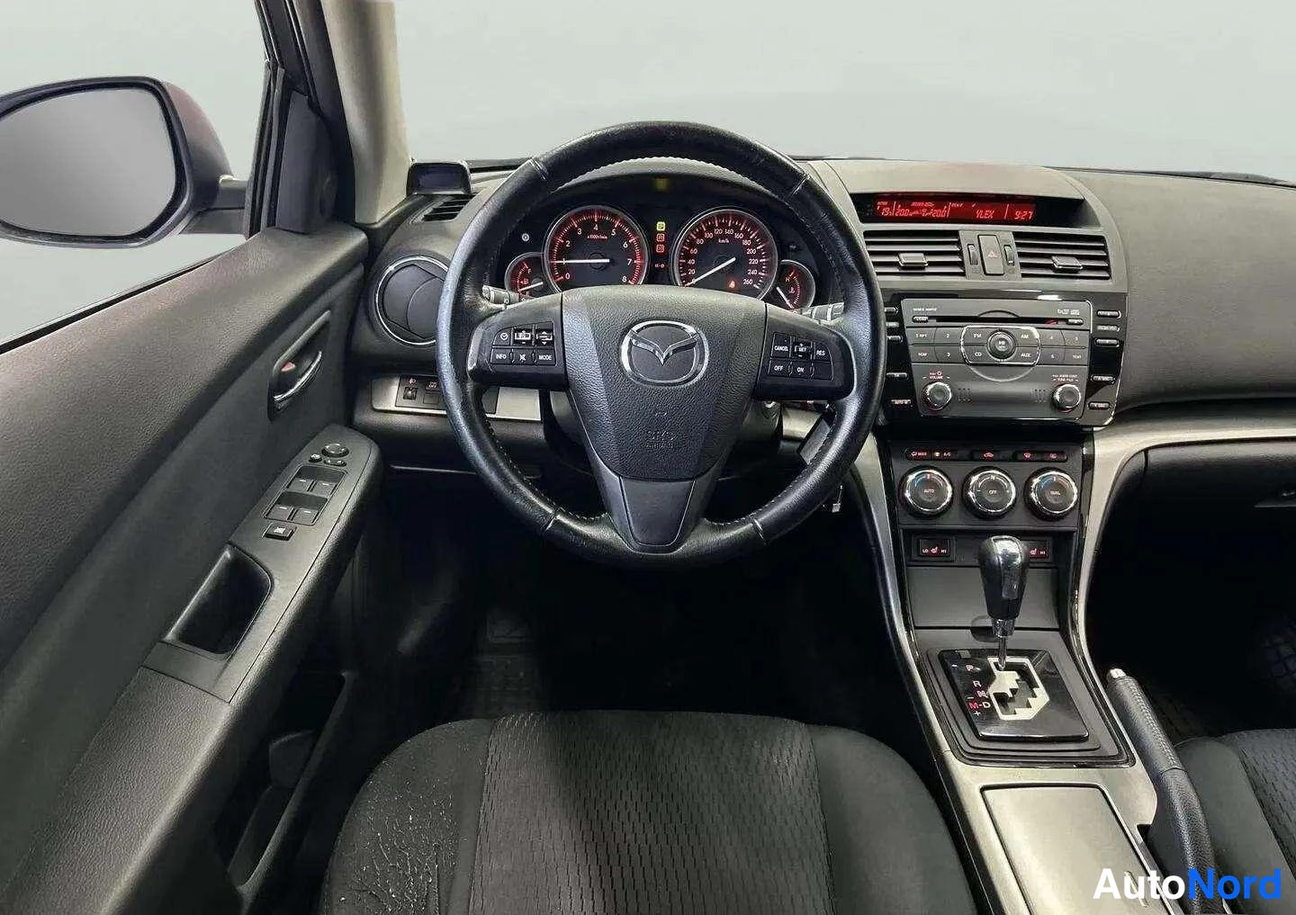 Yksityiskohta: Kuljettajan näkökulma ohjaamoon: Mazda-ohjauspyörä, analogiset mittarit, radio ja automaattivaihteen valitsin, vasen ovipaneeli näkyvissä.