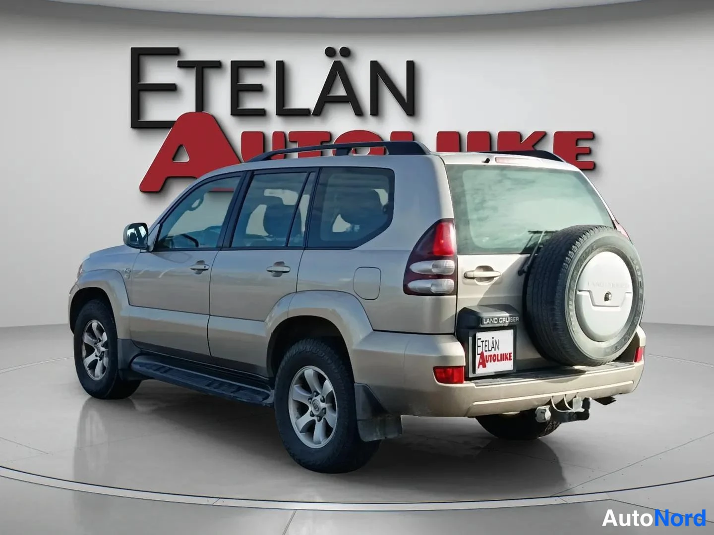 Takaviisto: Kuvakulma takaa ja vasemmalta sivulta: beige Toyota Land Cruiser -maasturu, jossa kattokaiteet, takana varapyörä ja vetokoukku, myyntihallin lattiassa.