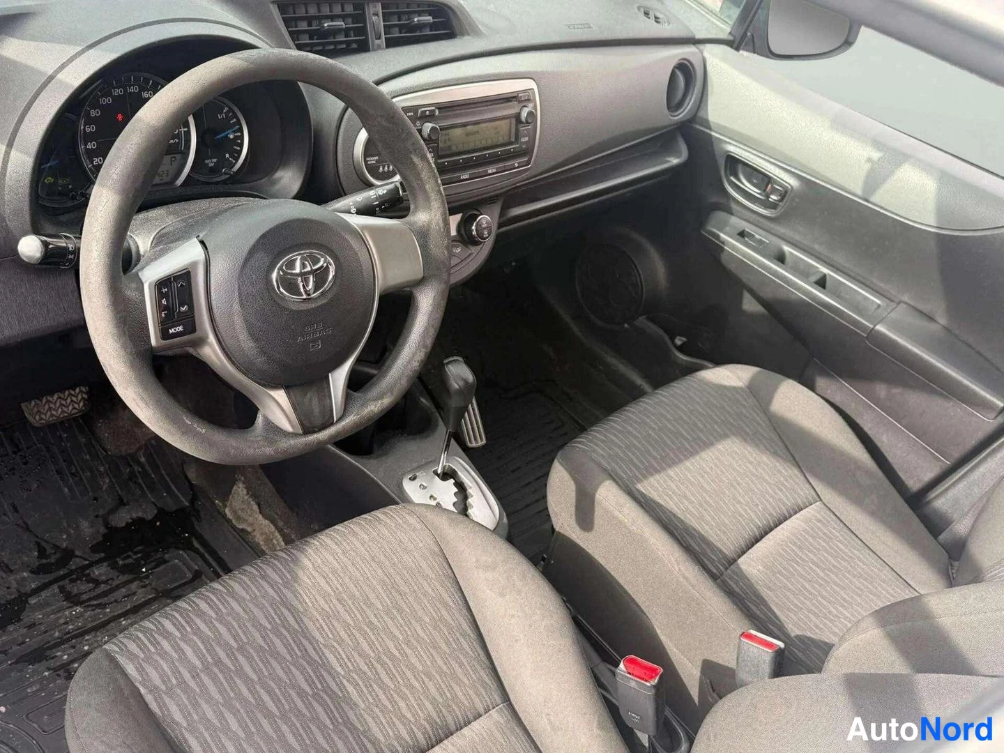 Muu: Toyota-henkilöauton ohjaamo: musta ohjauspyörä ja kojelauta, manuaali-ilmastointi (A/C), automaattivaihteiston valitsin ja kaksi tummaa kangasistuinta.