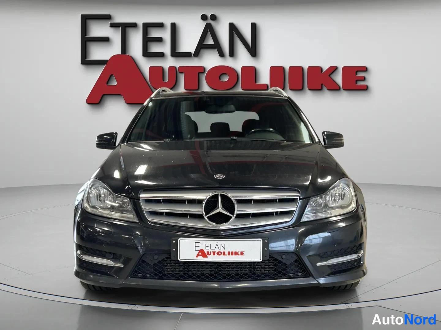 Edestä: Mustan Mercedes-Benz C-sarjan farmarin etunäkymä; Mercedes-tähtimerkki, kattokaiteet ja etupuskurin matala ilanottoaukko näkyvät suoraan edestä.