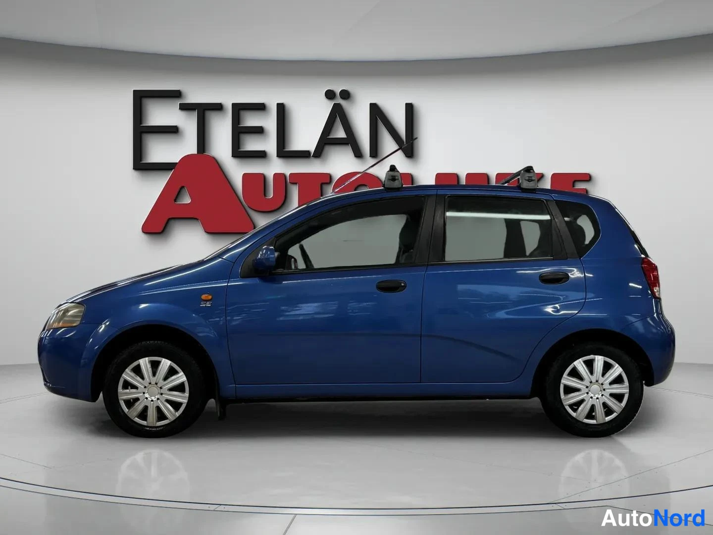Sivulta: Sinisen Chevrolet Kalos -henkilöauton sivunäkymä vasemmalta katsottuna, kattotelineet päällä.
