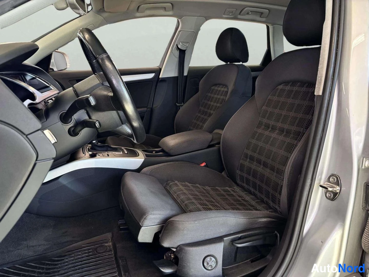 Muu: Audi A4:n etumatkustamointi: mustat kangasistuimet kuviollisilla istuinkankailla, ohjauspyörä ja keskikonsoli, kuva otettu avatusta oikeasta takaovesta.