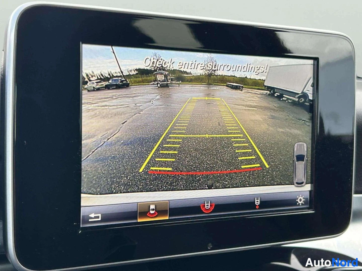 Muu: Mercedes-Benzin keskinäyttö näyttää takakameran kuvan keltaisine kaistaneuvoineen ja tekstin 'Check entire surroundings!'.