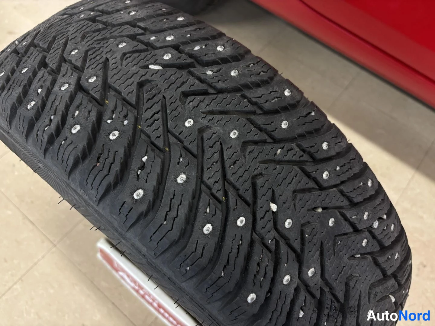 Yksityiskohta: Detail shot of a studded winter tyre; red car body panel visible in the background.