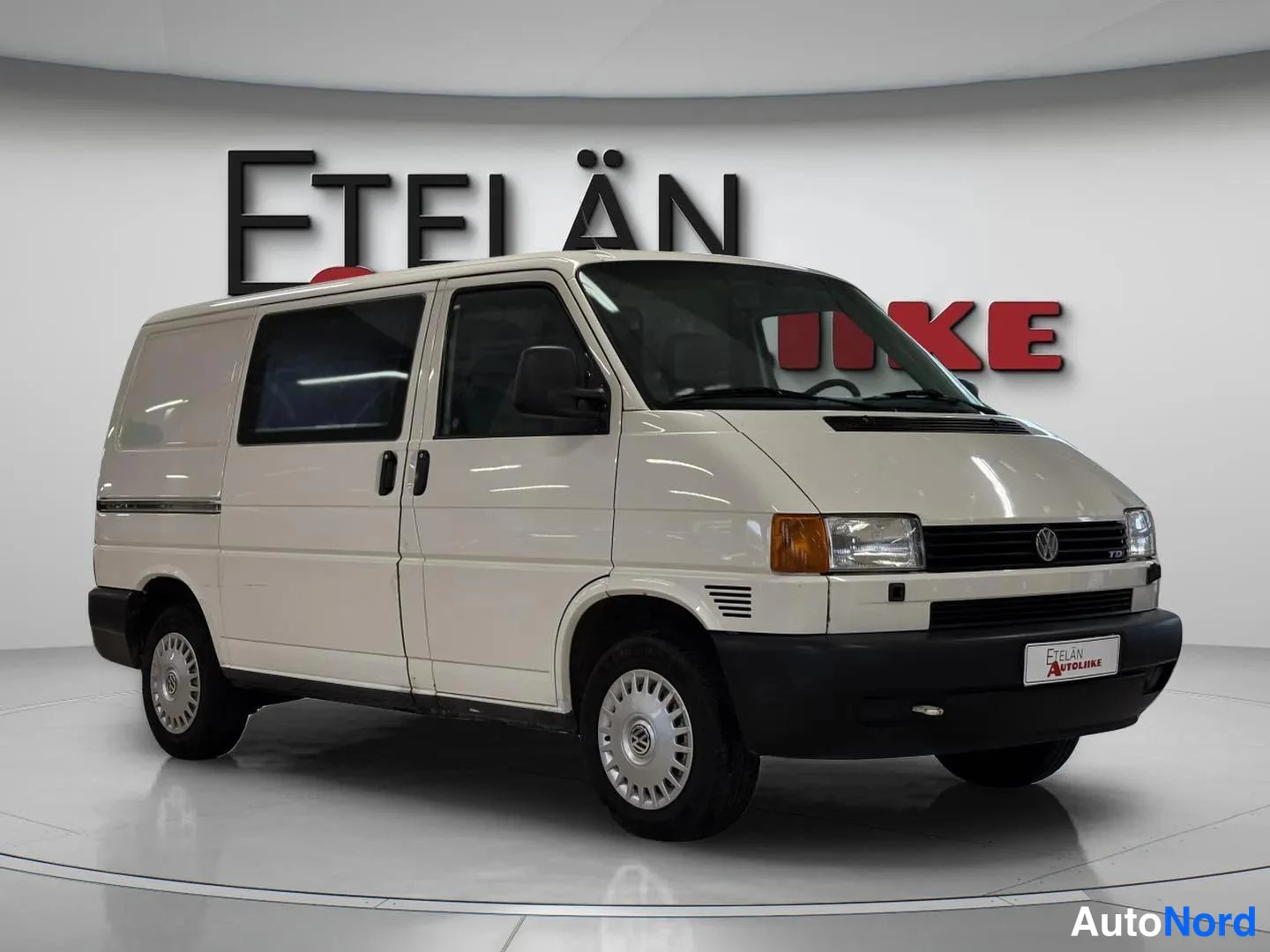 Etuviisto, nokka oikealle: Valkoinen Volkswagen Transporter -pakettiauto nähty edestä oikealta, myyjän tilassa valkoista taustaa vasten.