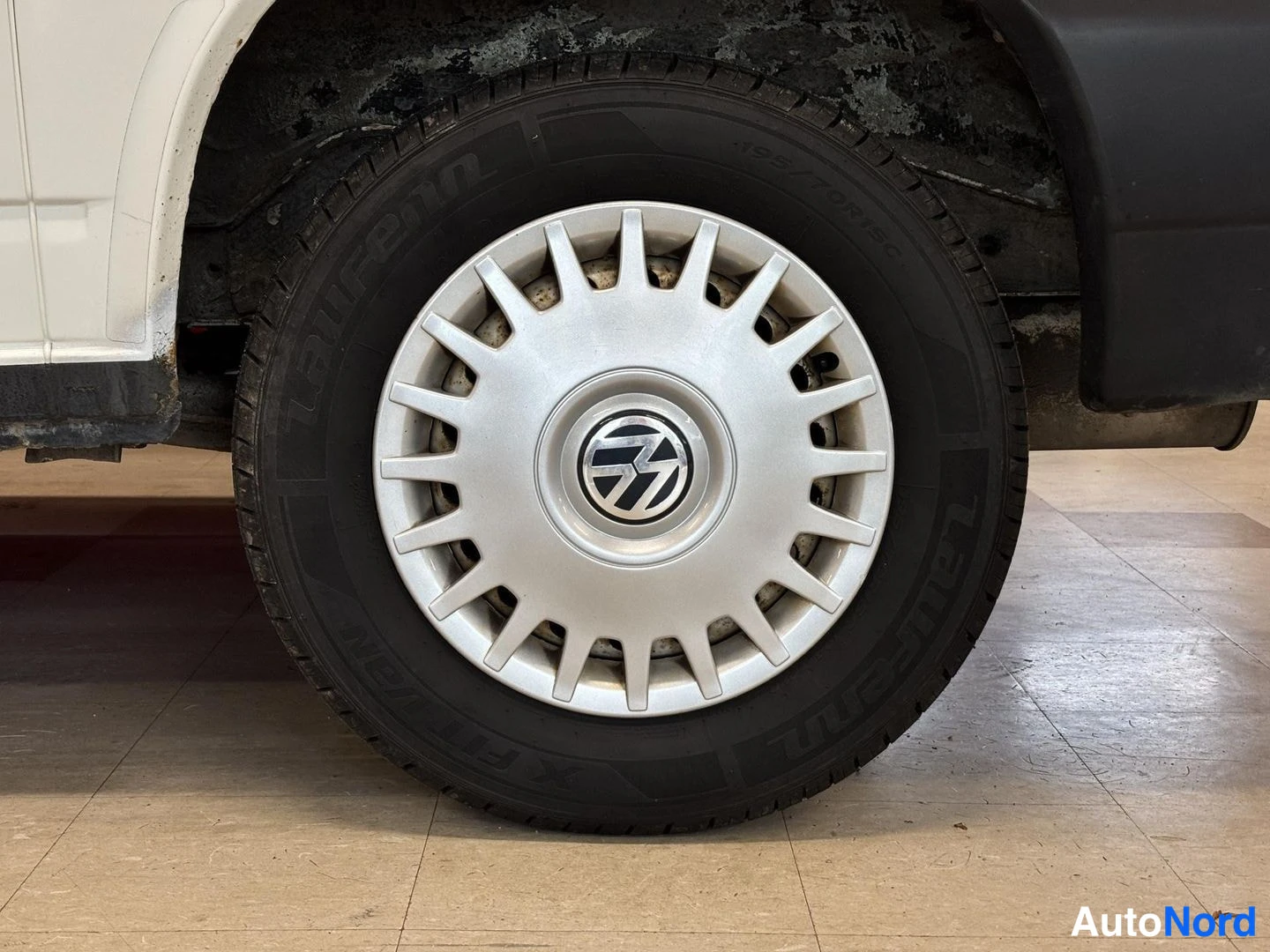 Yksityiskohta: Kuva vanhemman mallisesta hopeanvärisestä pyöränmaskista, jossa on musta Volkswagen-tunnus keskellä. Maskin pinnassa näkyy pinttynyttä likaa, rengas on musta.