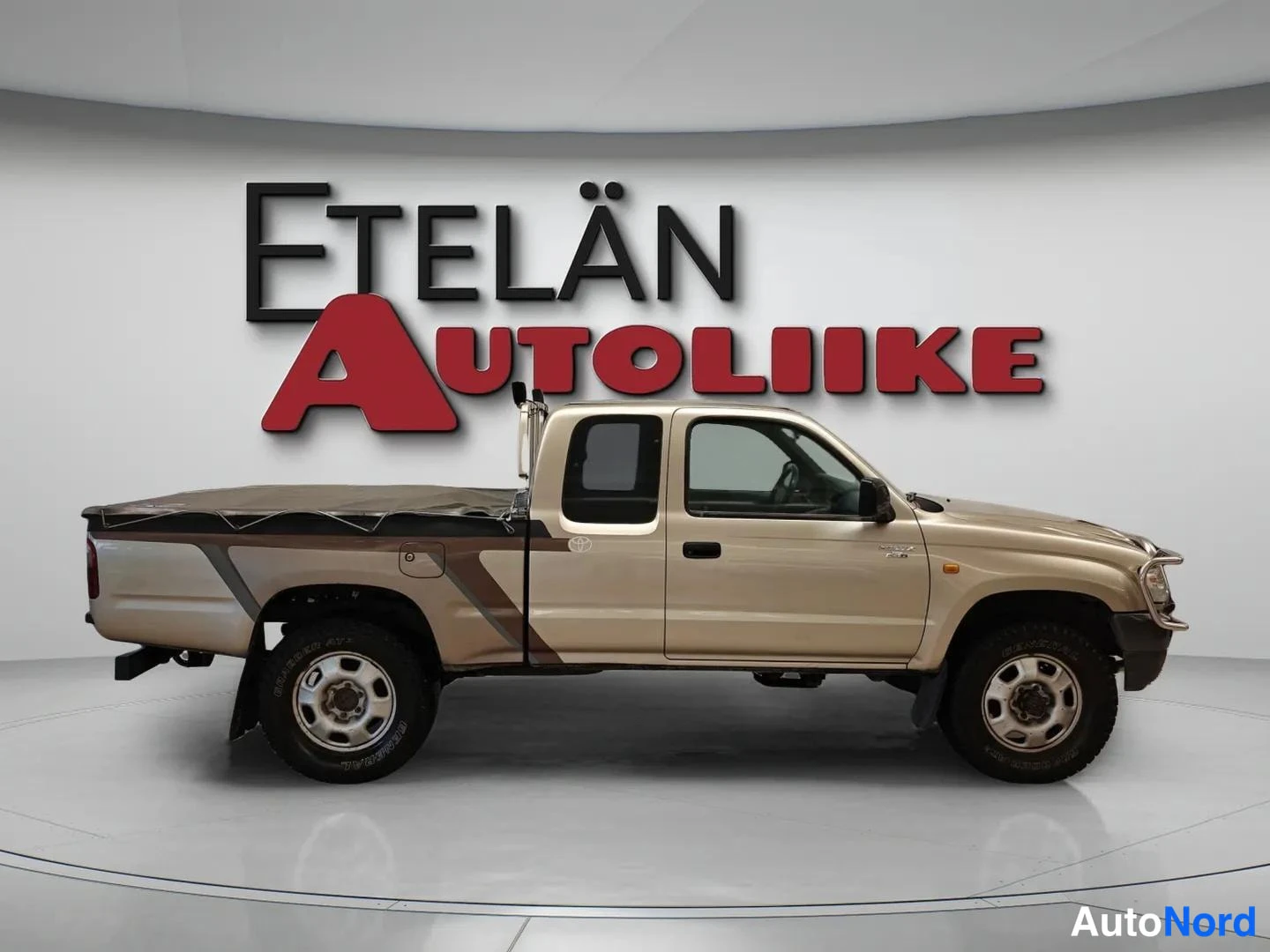 Sivulta: Auton sivuprofiili, beige Toyota-pickup kaksiovisella ohjaamolla ja peitetyllä lavaosalla.