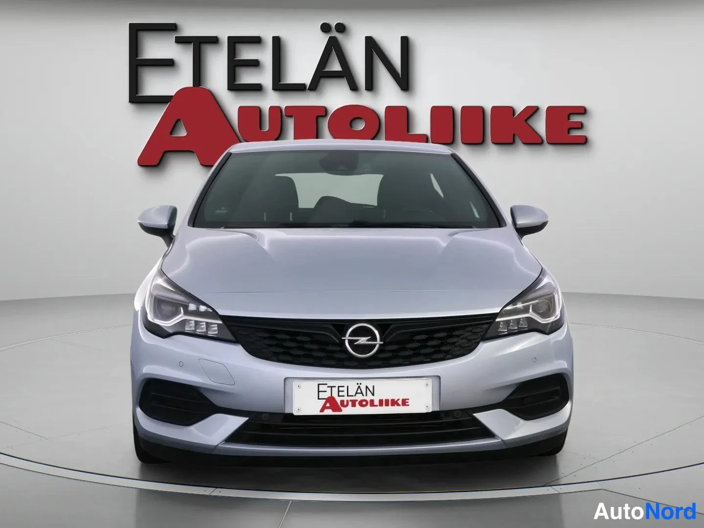 Edestä: Auton etusivu: hopeanvärinen viisiovinen tila-automallin etupää, Opel-merkki etusäleikössä, myyjän logo taustalla.