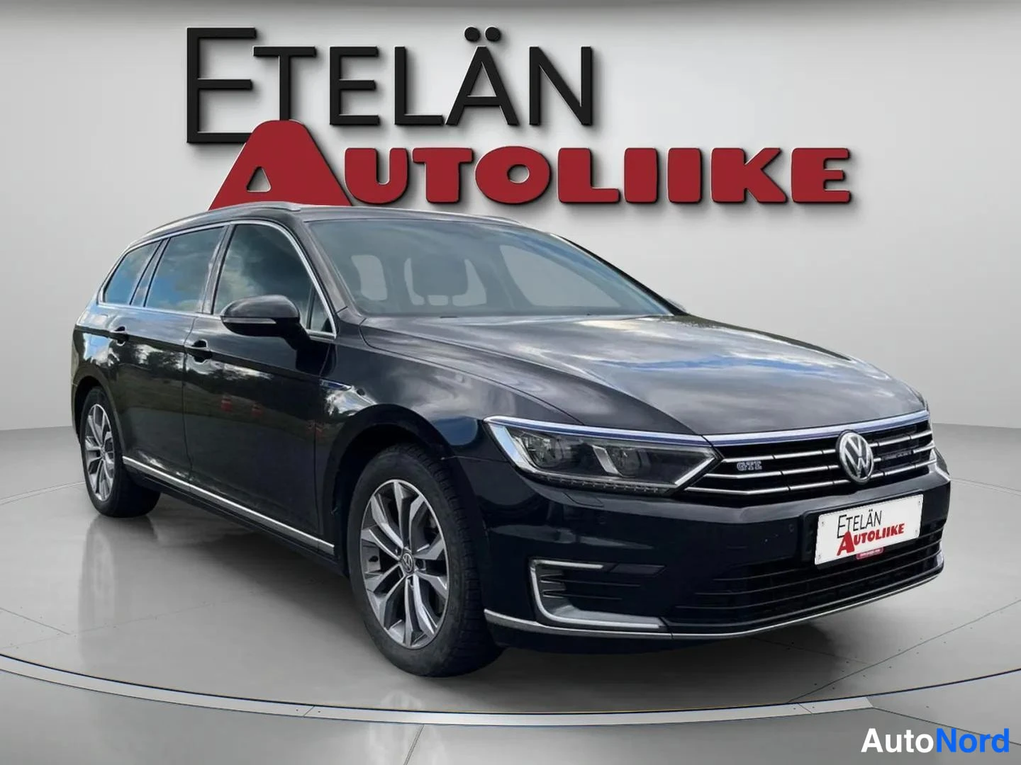 Etuviisto, nokka oikealle: Tummansininen farmariauto etu- ja sivukulmassa autonäyttelytaustassa