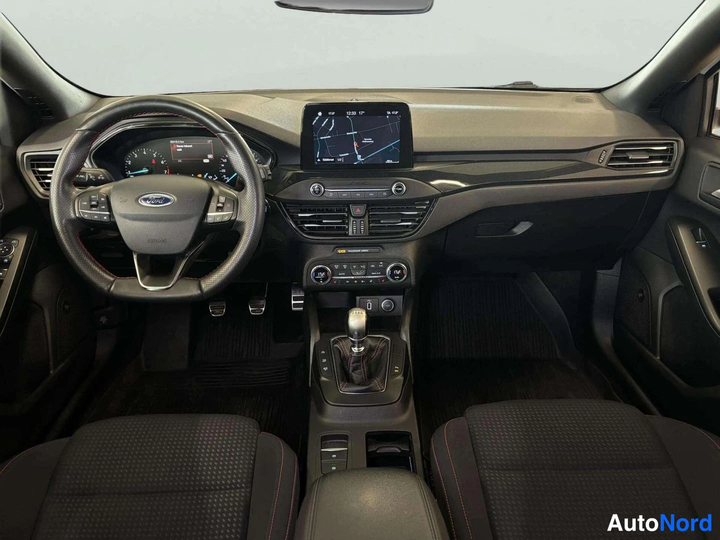 Muu: Auton kojelauta edestäpäin: Ford-ohjauspyörä, kartta näyttävä keskinäyttö ja manuaalivaihdekeppi.