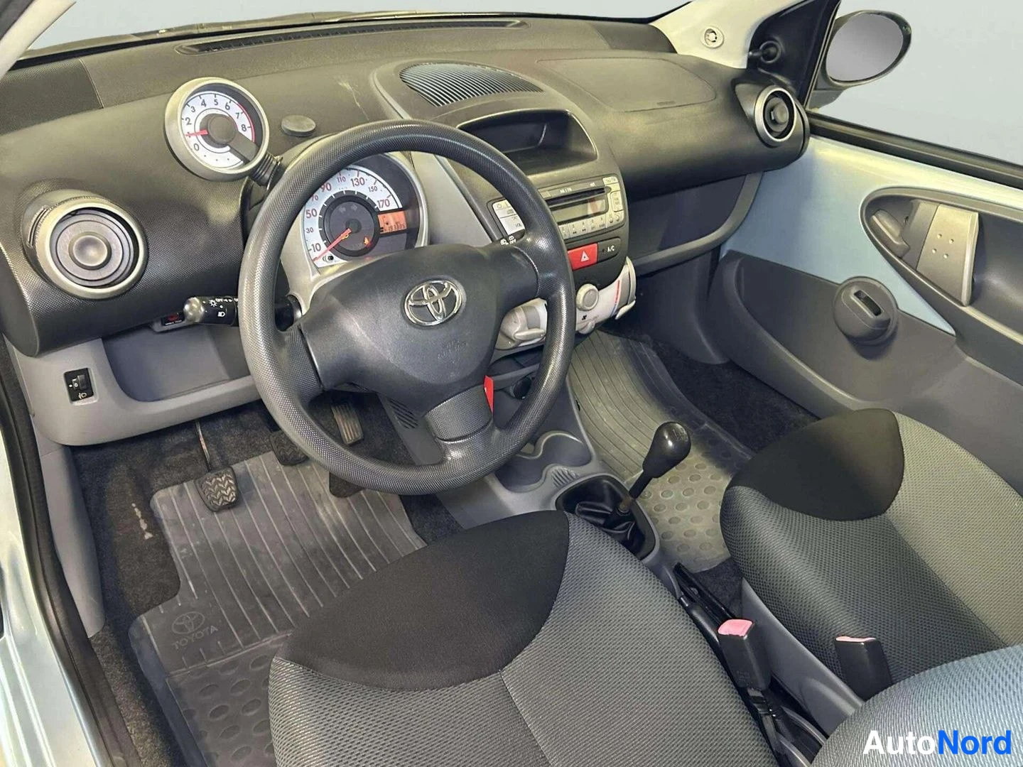 Muu: Ohjaamo kuvattuna kuljettajan puolelta: Toyota-ratti, analogiset mittarit, manuaalivaihteiston vaihdekeppi ja tummat penkit.