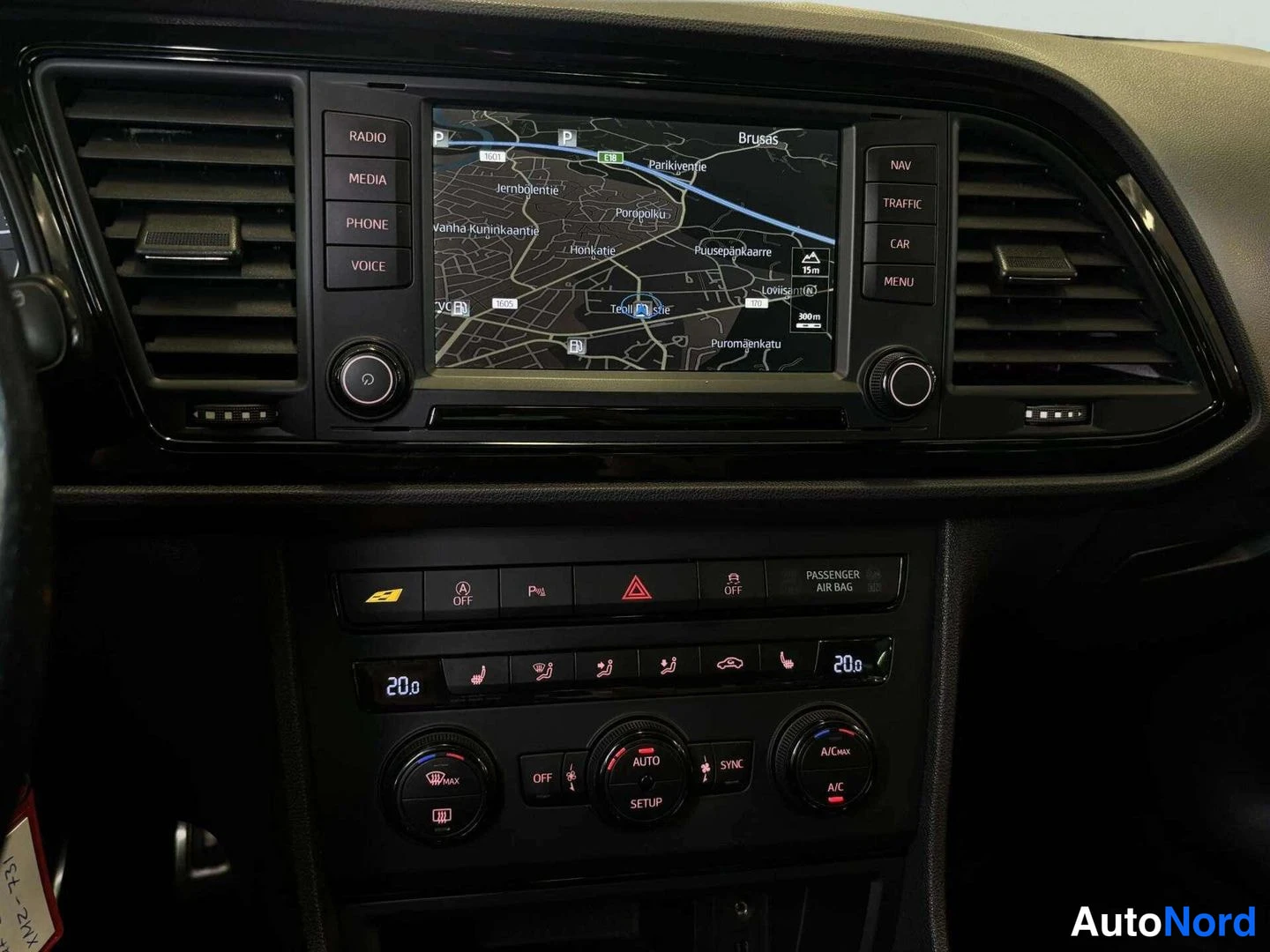 Ohjaamo: Dashboard showing navigation screen displaying city map and route guidance, dual-zone climate control panel below it, hazard light button active.