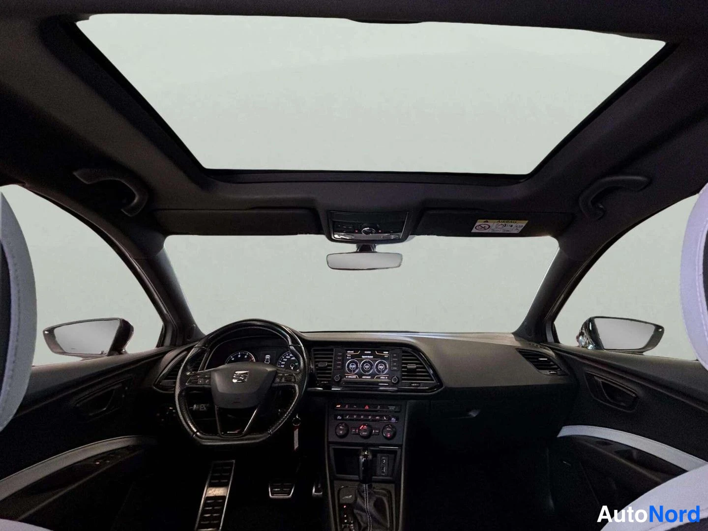 Ohjaamo: Wide interior shot looking through the front windshield. A large glass sunroof dominates the ceiling area. Steering wheel with leather trim is visible below, fl