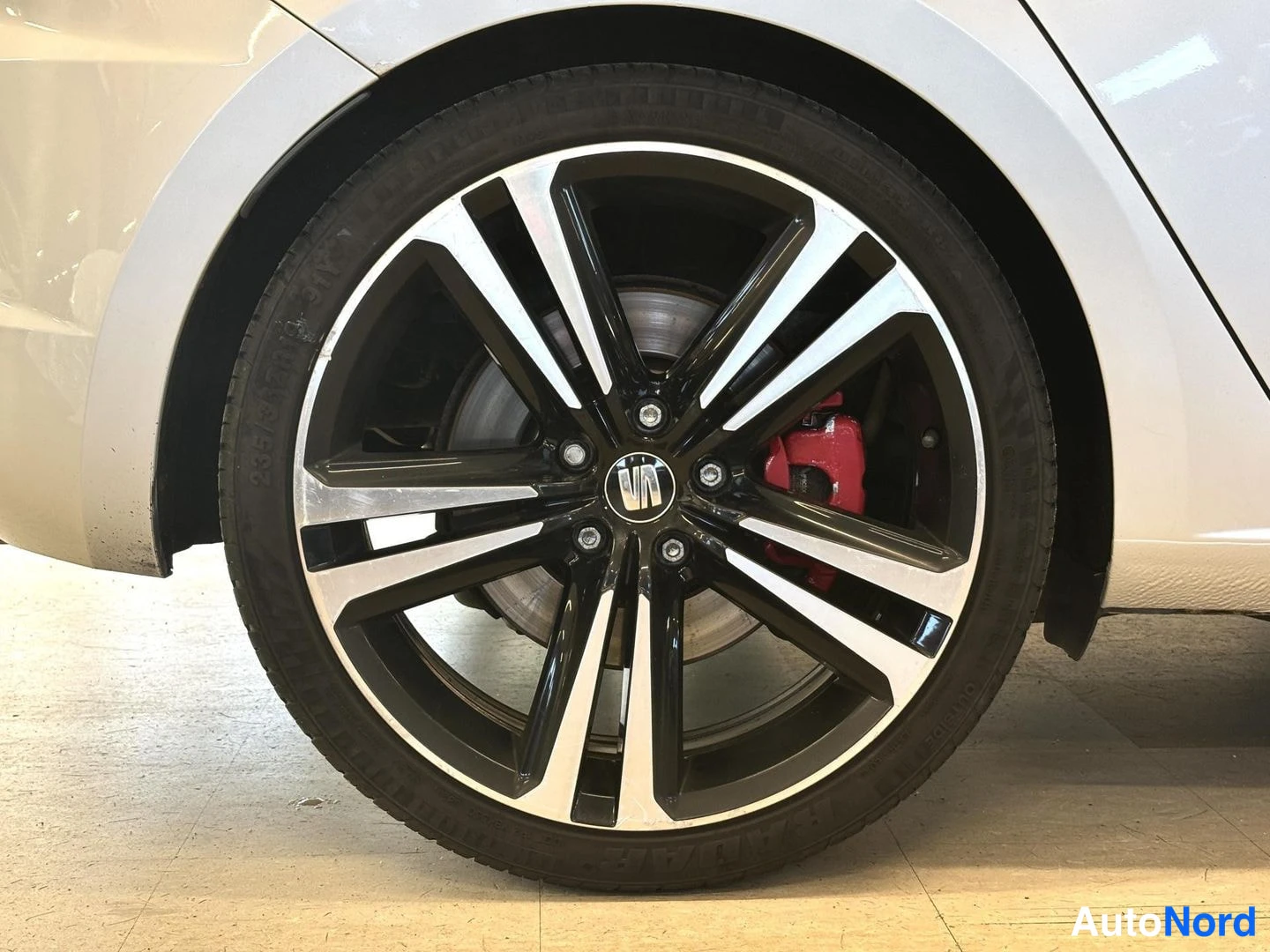 Yksityiskohta: Close-up of a two-tone alloy wheel with a multi-spoke design in glossy black and machined silver. Behind the spokes, a red brake caliper is visible mounted to a
