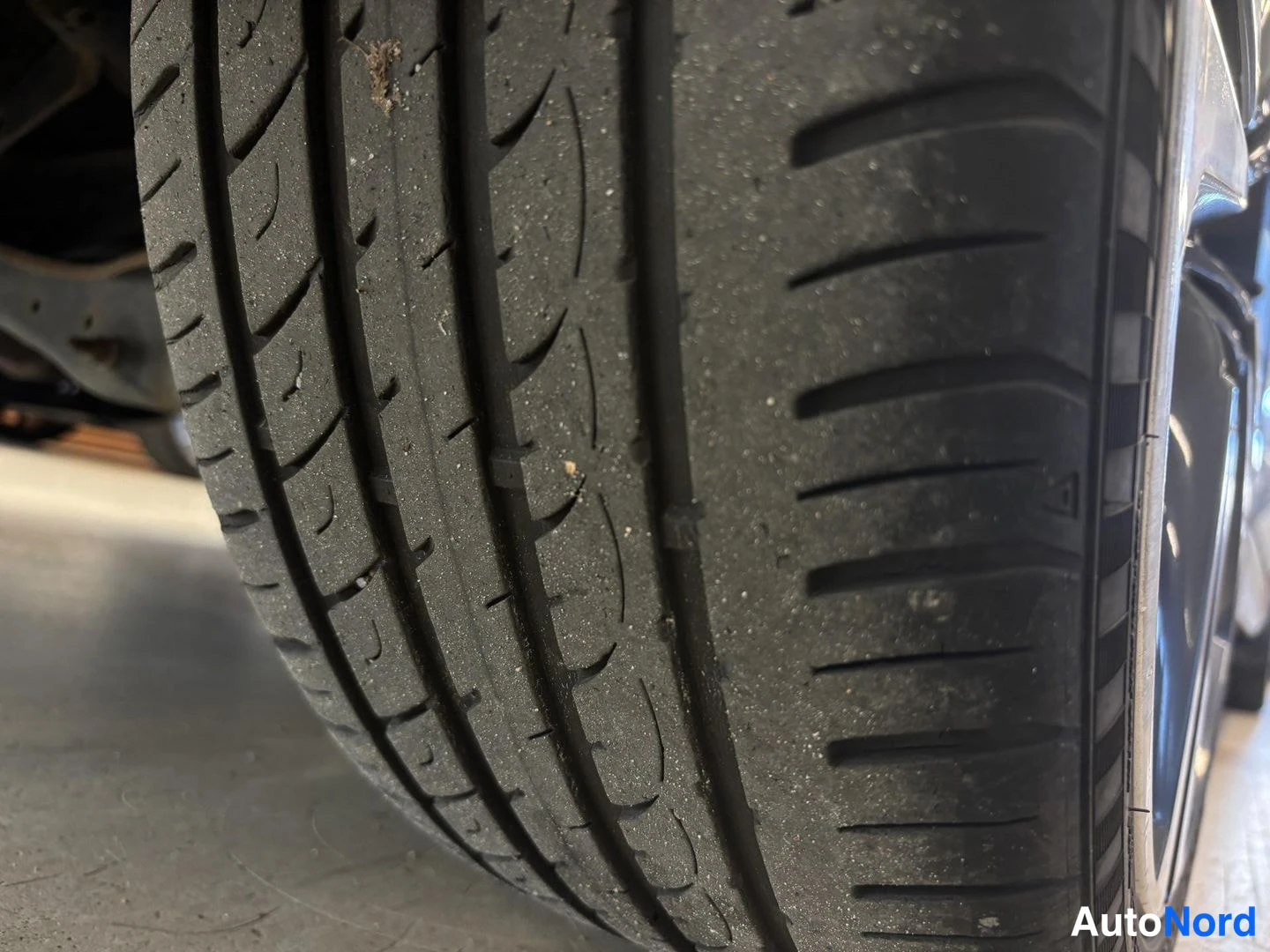 Yksityiskohta: Close-up view of a summer tyre's tread surface showing circumferential grooves and sipes. Fine sand or grit particles are embedded in the rubber. A portion of a
