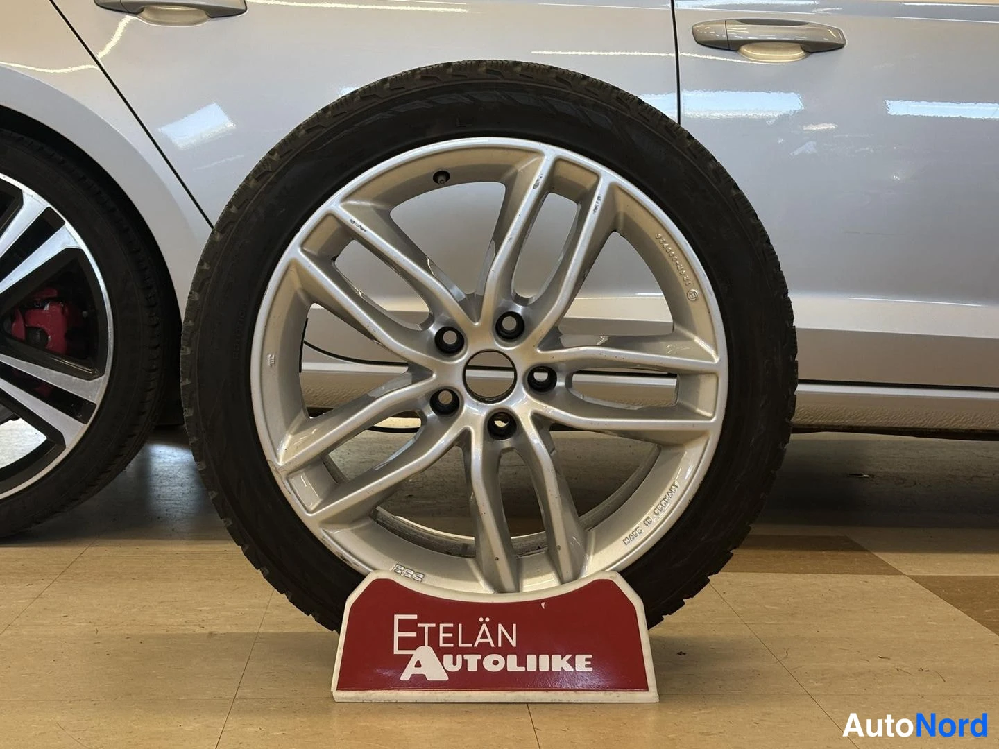 Yksityiskohta: A silver five-twin-spoke alloy wheel fitted with a dark tread tyre standing upright on a display stand that reads 'Etelän Autoliike'. The white car flank and a