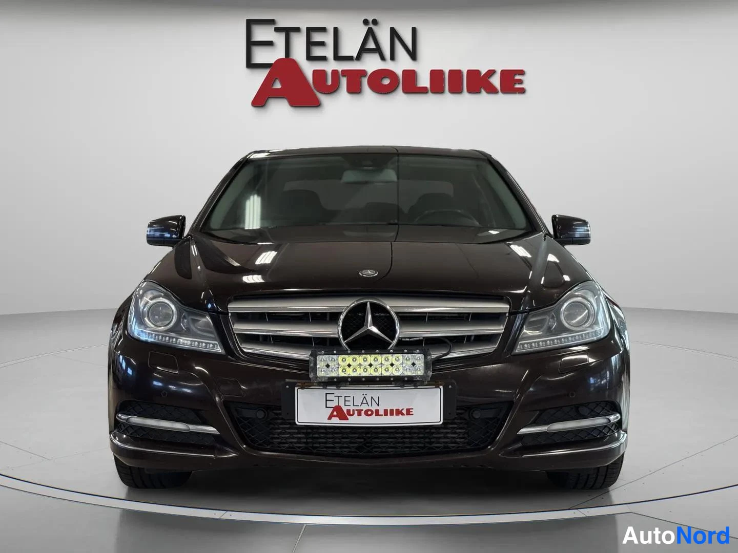 Edestä: Etukuvassa Mercedes-Benz C-sedan; etusäleikkö Mercedes-tähdellä, isot ajovalot ja etupuskuri täyttävät kuvan. Etupuskurissa kiinnitetty lisävalokiskot.