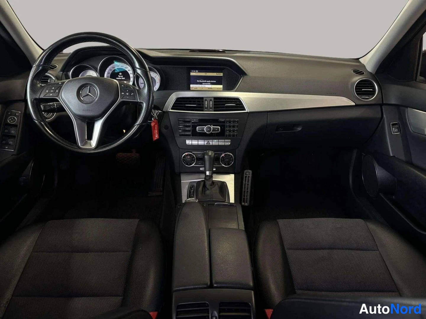 Edestä: Ohjaamo suoraan edestä: Mercedes-ohjauspyörä ja mittaristo, keskusnäyttö Bluetooth-viestillä, automaattivaihteen valitsin ja kaksi etuistuinta.