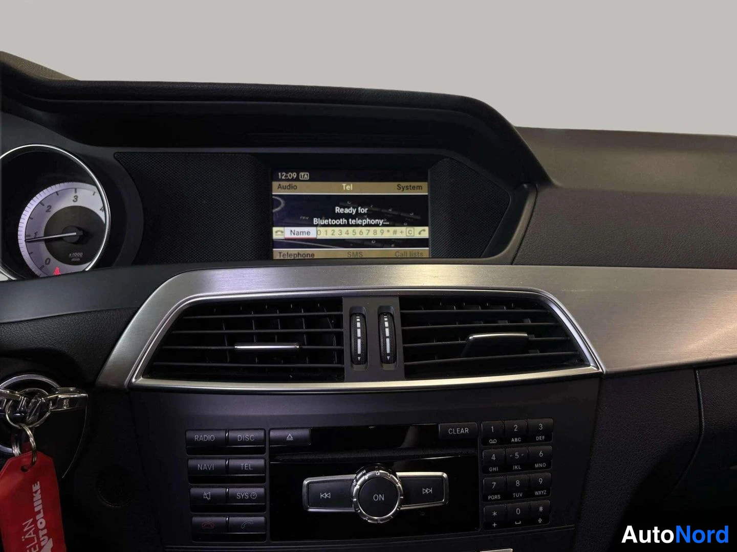 Edestä: Kojelaudan näyttö näyttää tekstiä 'Ready for Bluetooth telephony' ja näppäimistön, alla radio/navi/tele-napit ja ilmastointilaitteet. Avaimet ovessa.