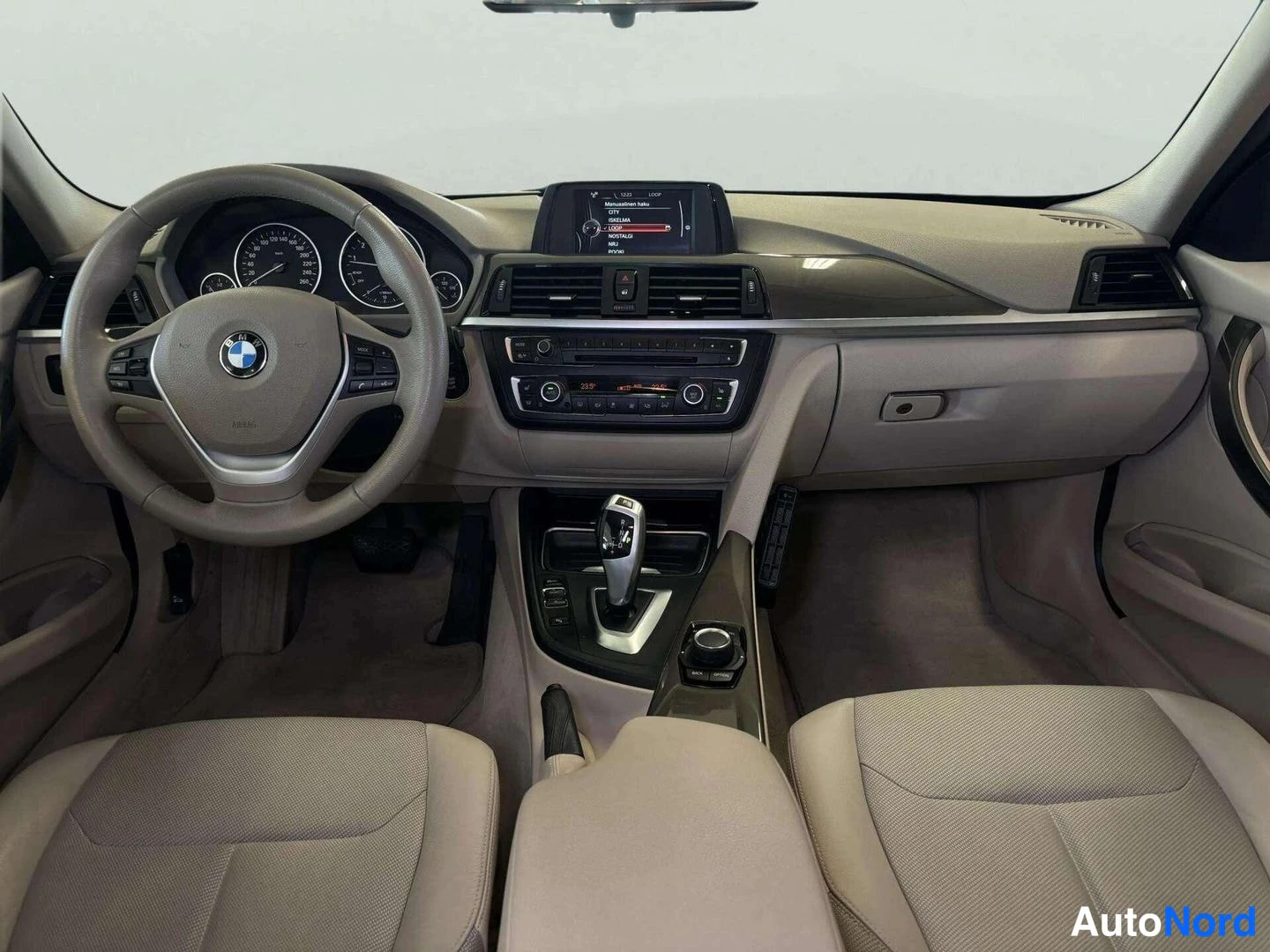 Edestä: Auton kojelauta suoraan edestä: BMW-ohjauspyörä, mittarit, keskikonsolin näyttö ja automaattivaihteen valitsin. Etuistuimet ovat vaaleaa beigetä.