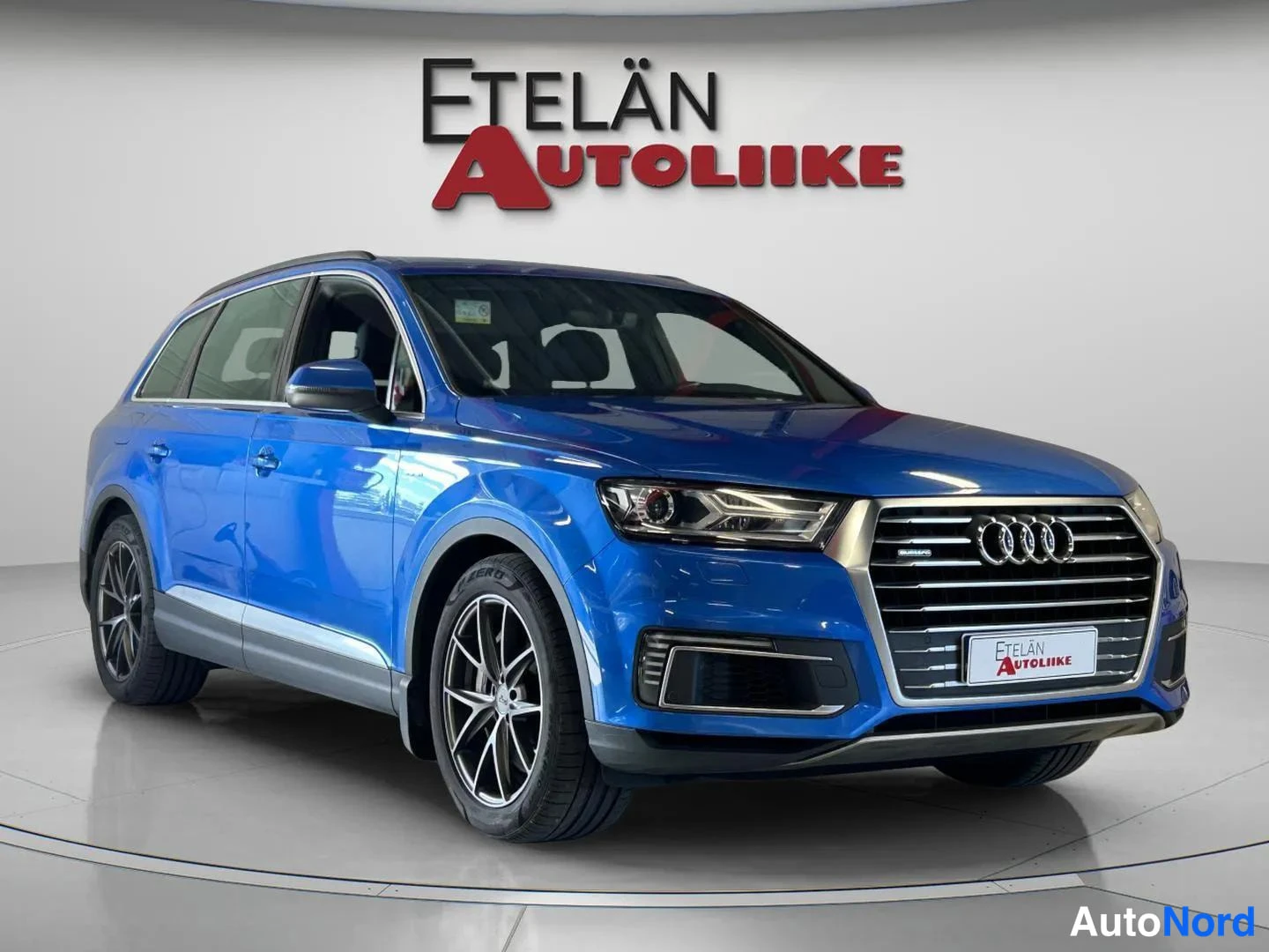 Etuviisto, nokka oikealle: Sininen Audi Q7 etukulmasta, keulagrilli ja vasen kylki näkyvissä studiotaustaa vasten.