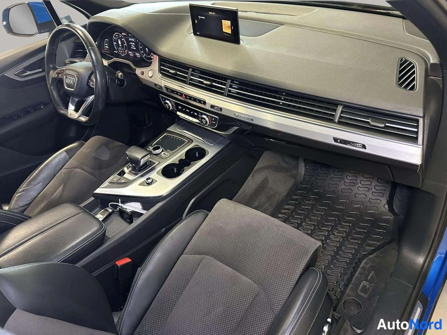 Edestä: Audi Q7:n sisätilat matkustajan puolelta kuvattuna: kojelauta, vaihteenvalitsin, kuppitelineet ja kumimatot näkyvissä.