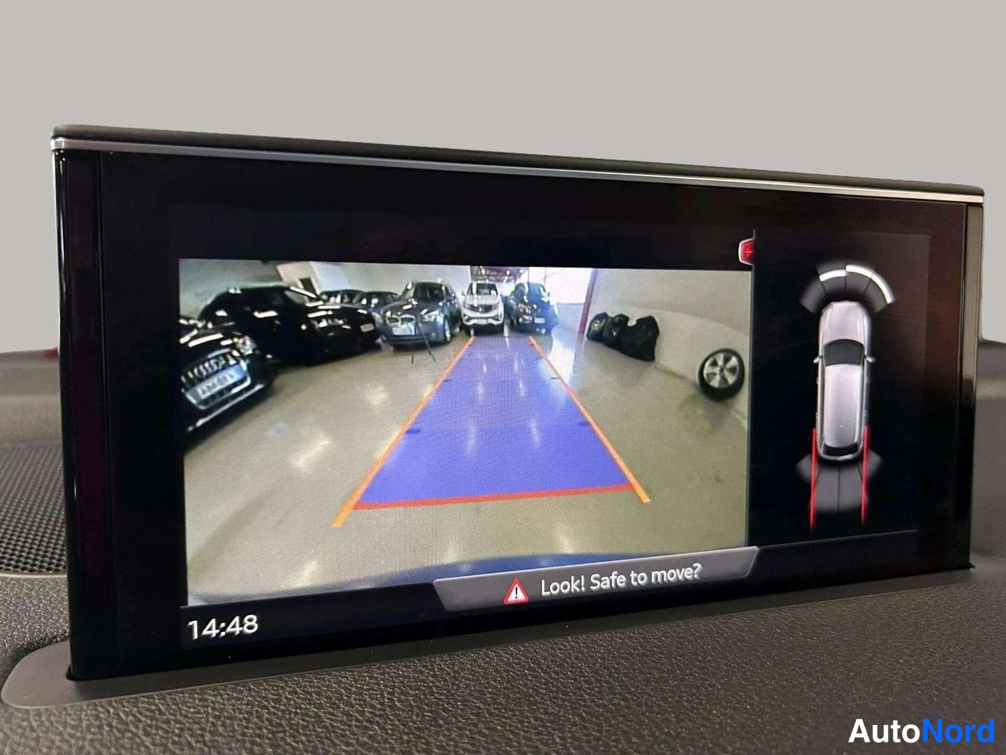 Yksityiskohta: Audi Q7:n infotainment-näyttö näyttää peruutuskameran kuvan ja kaarivalvontagrafiikan tekstillä 'Look! Safe to move?'.