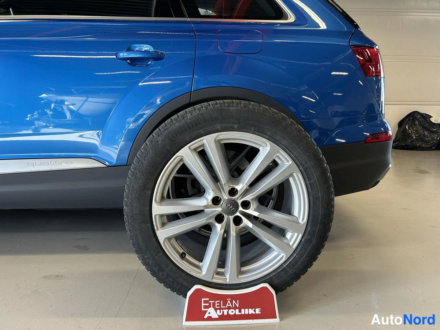 Takaviisto: Sinisen Audi Q7:n takapyörä ja hopea kevytmetallivanne lähikuvassa, kyljessä näkyy quattro-merkintä. Auton alla on Etelän Autoliike-teline.
