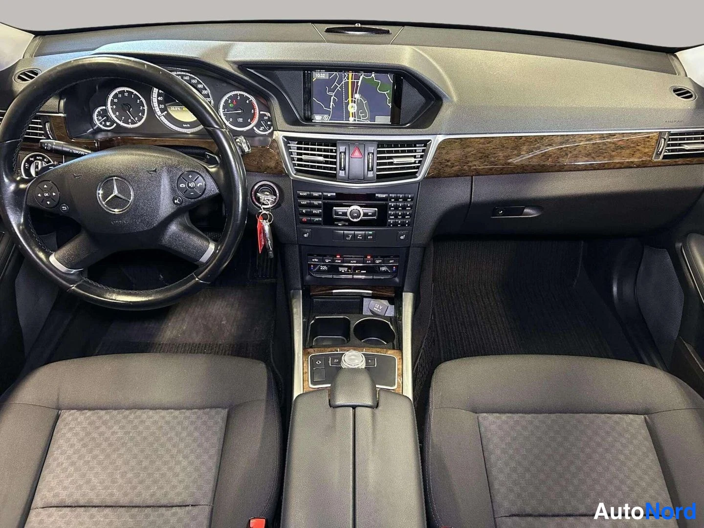 Yksityiskohta: Mercedes-Benzin ohjaamo etualalta kuvattuna: ratissa Mercedes-merkki, kojelaudalla navigointinäyttö ja puiset koristelistat, keskellä pyörien lämmityssäätimet j