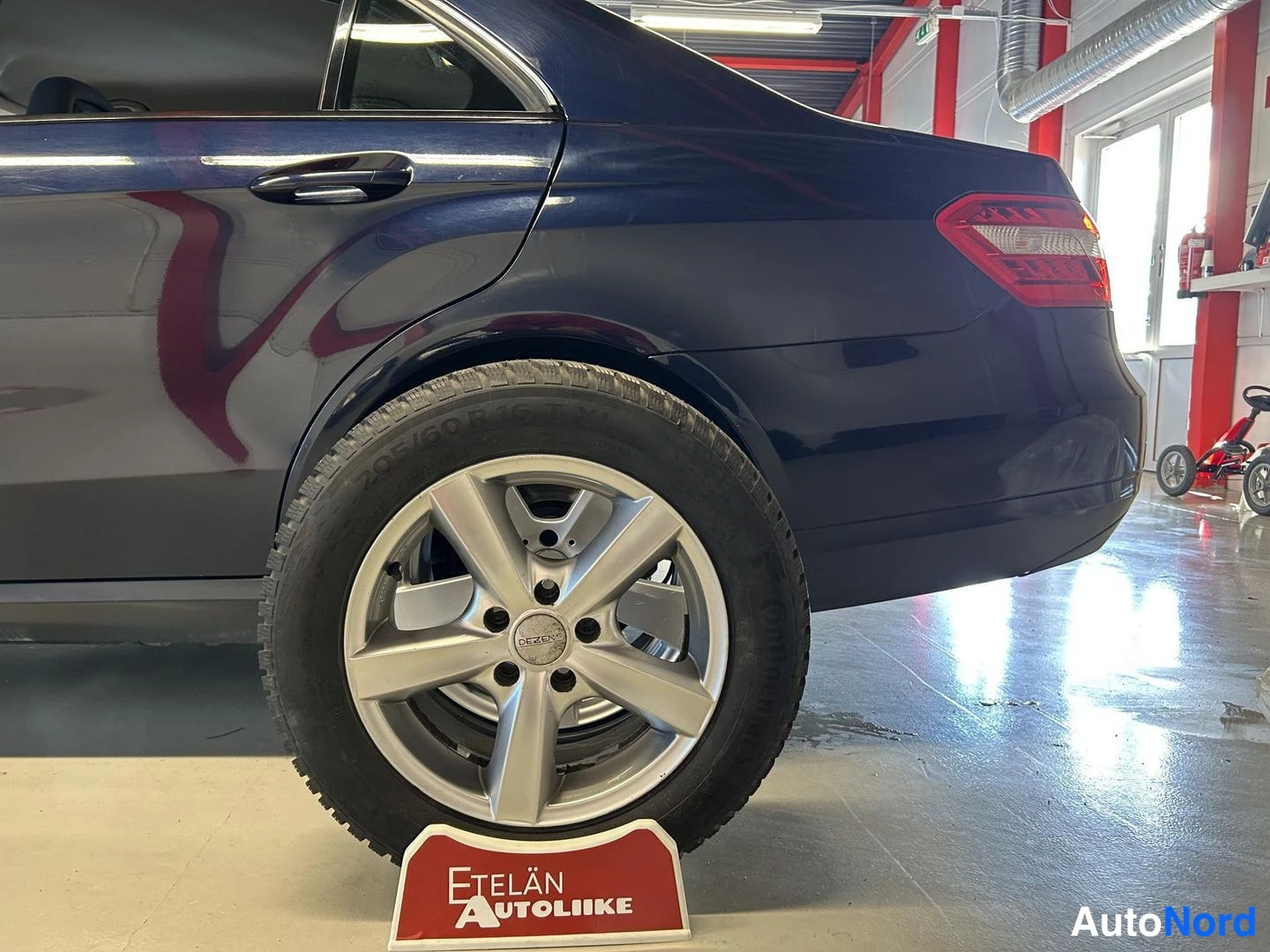Takaviisto: Tummansinisen auton takakulma ja takapyörä hopeisella Mercedes-vanteella; alla punainen Etelän Autoliike -kyltti.
