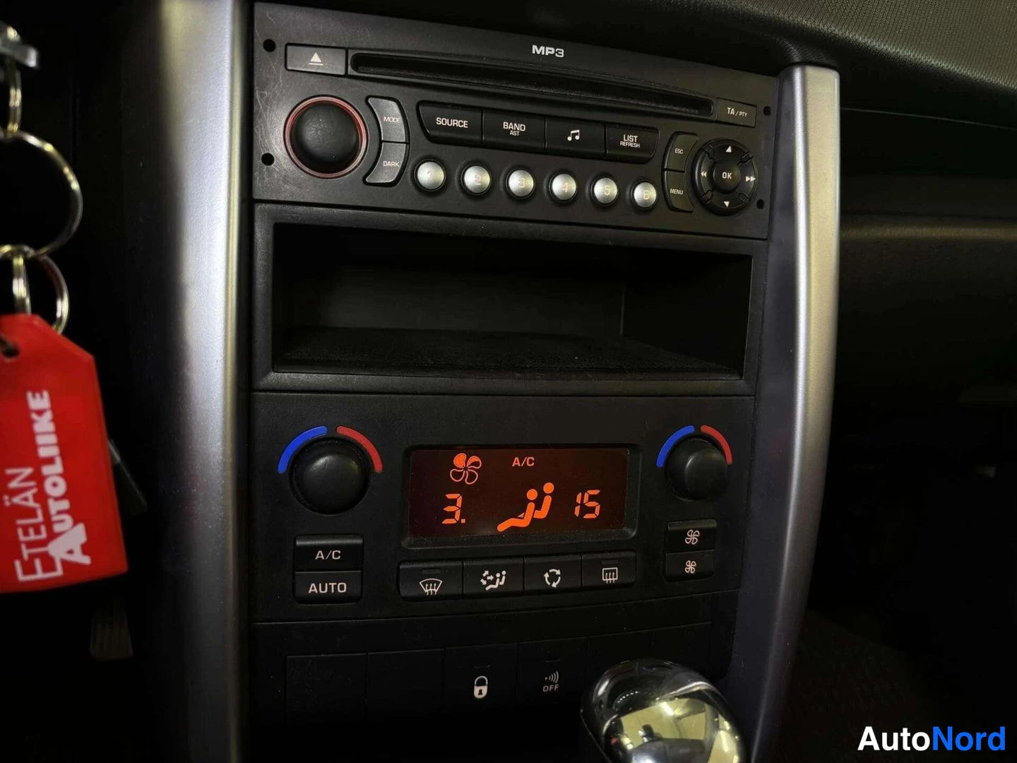 Muu: Keskikonsolin MP3-radio-CD-yksikkö lähikuvassa, alla ilmastointipaneeli A/C- ja AUTO-painikkeilla ja oranssilla näytöllä, vasemmalla punainen Etelän Autoliike -